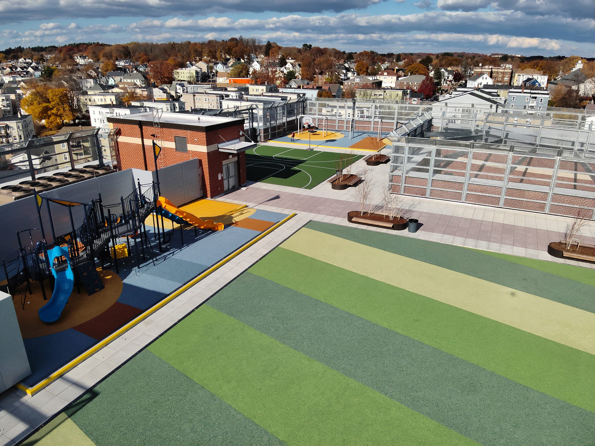 Aerial view of a modern rooftop with a playground, sports court, green spaces, and a city skyline background.