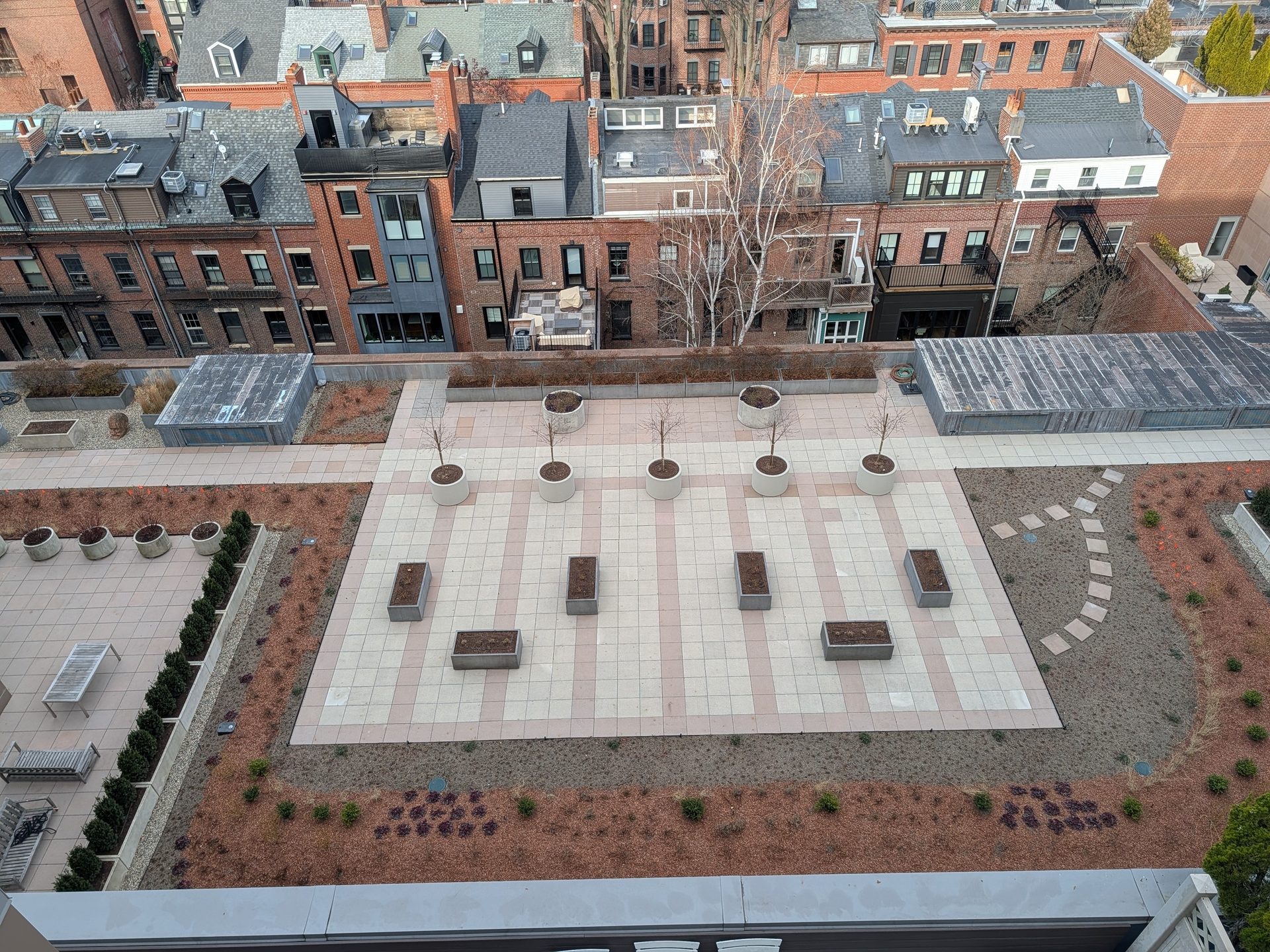 Aerial view of a rooftop garden with patterned tiles, planters, trees, and residential buildings.