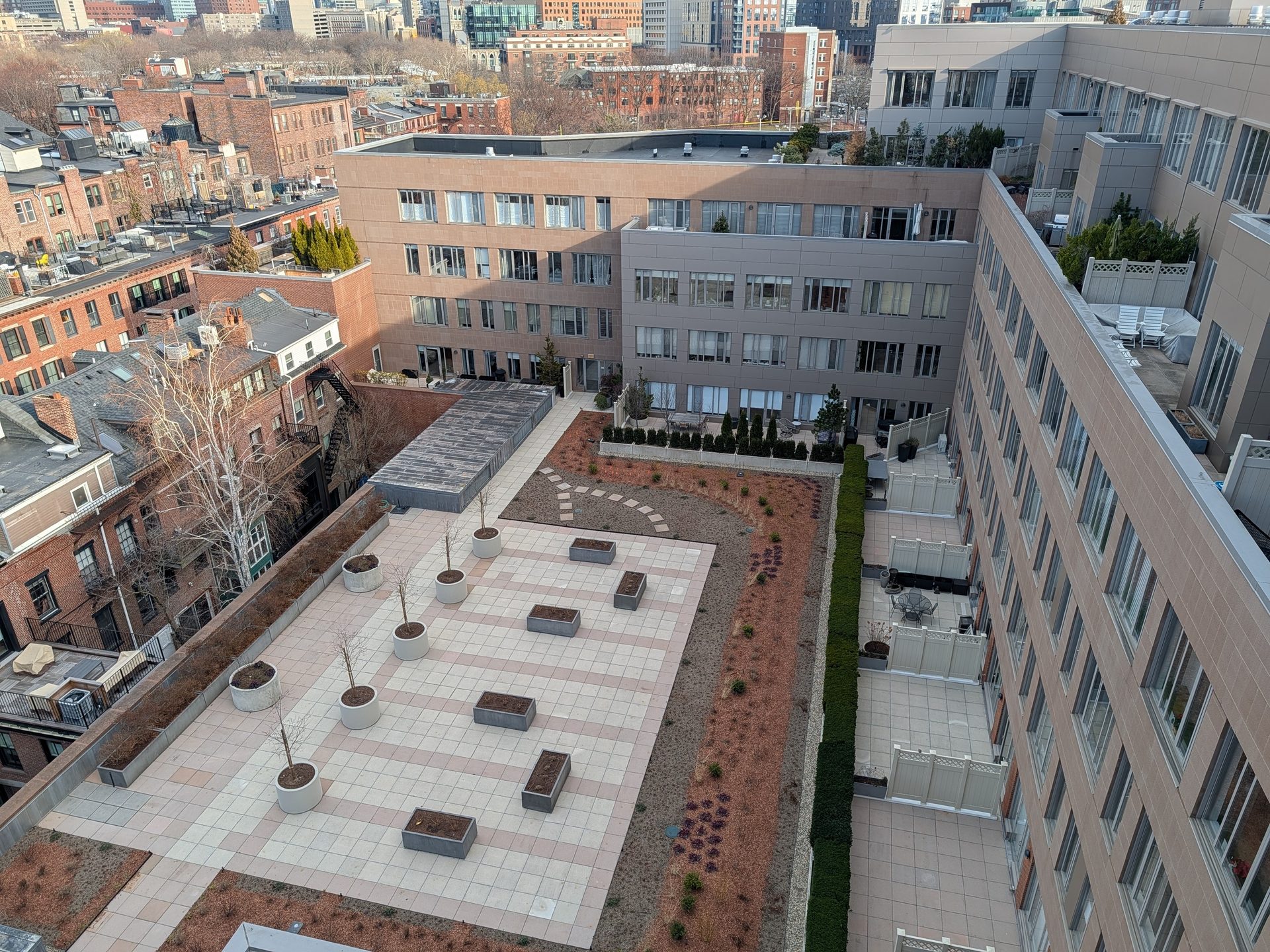 Aerial view of a city rooftop garden with tiled paths, planters, small trees, and surrounding apartment buildings.