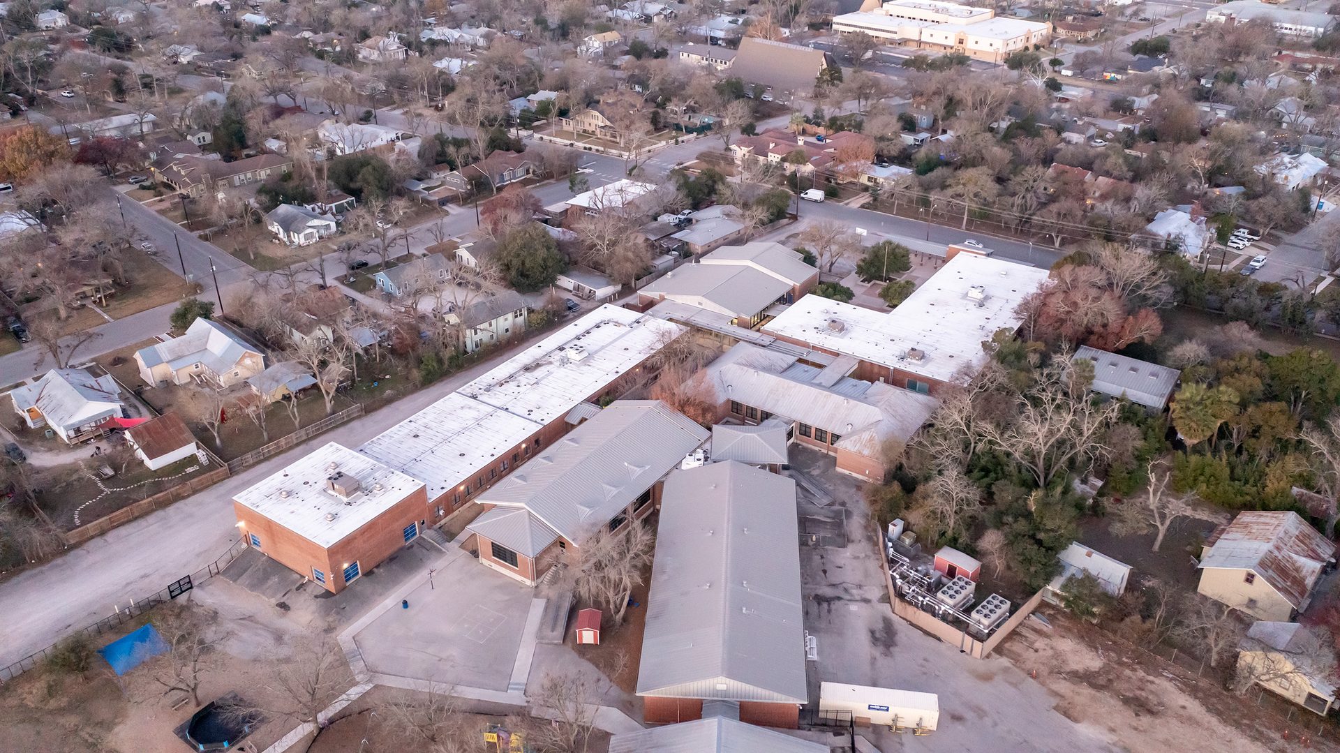 Aerial view of a school complex with light-colored roofs, surrounded by a residential neighborhood and bare trees.