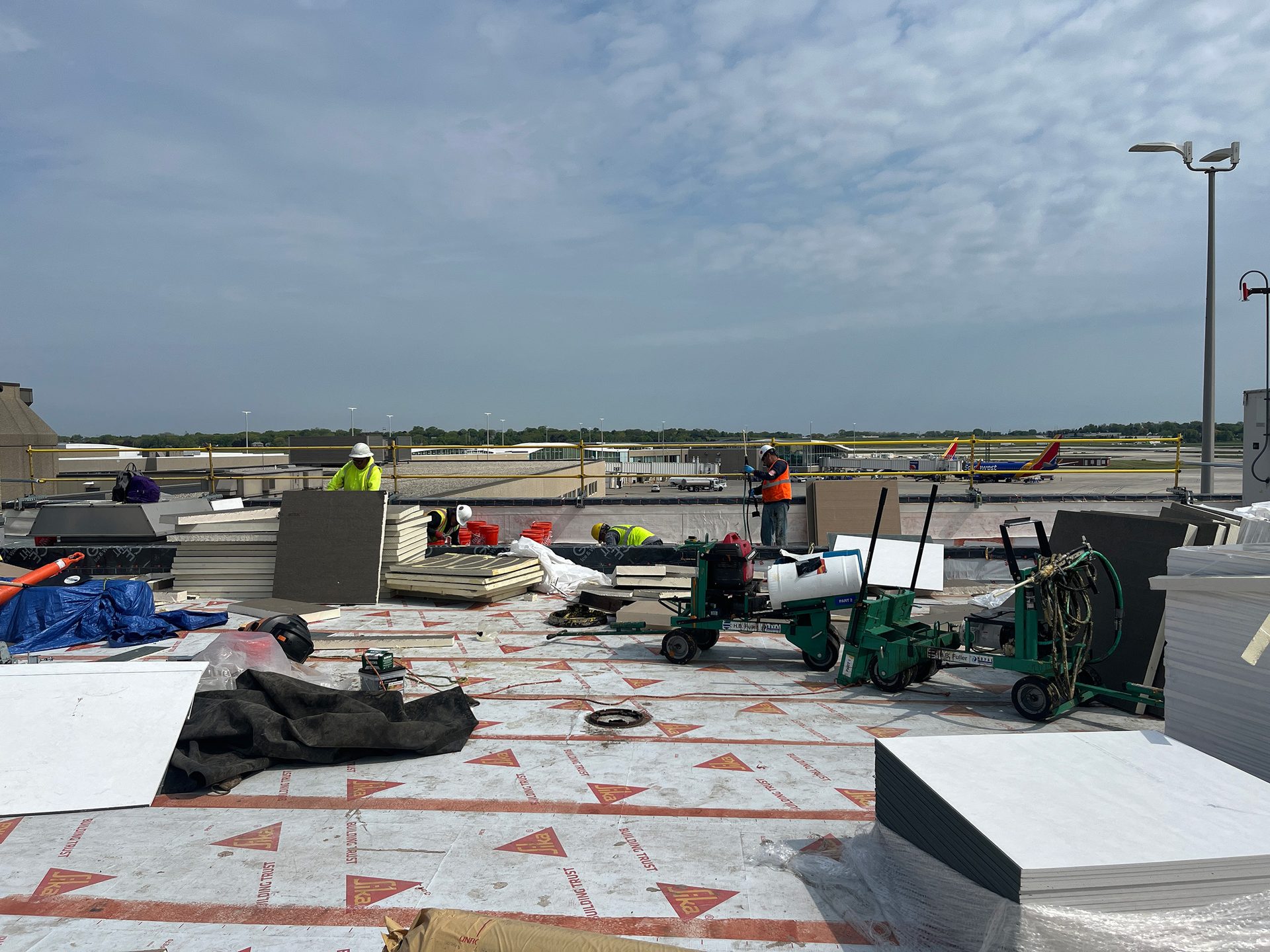 Construction workers on a large rooftop, surrounded by building materials and equipment, with an airport tarmac and airplanes in the background.