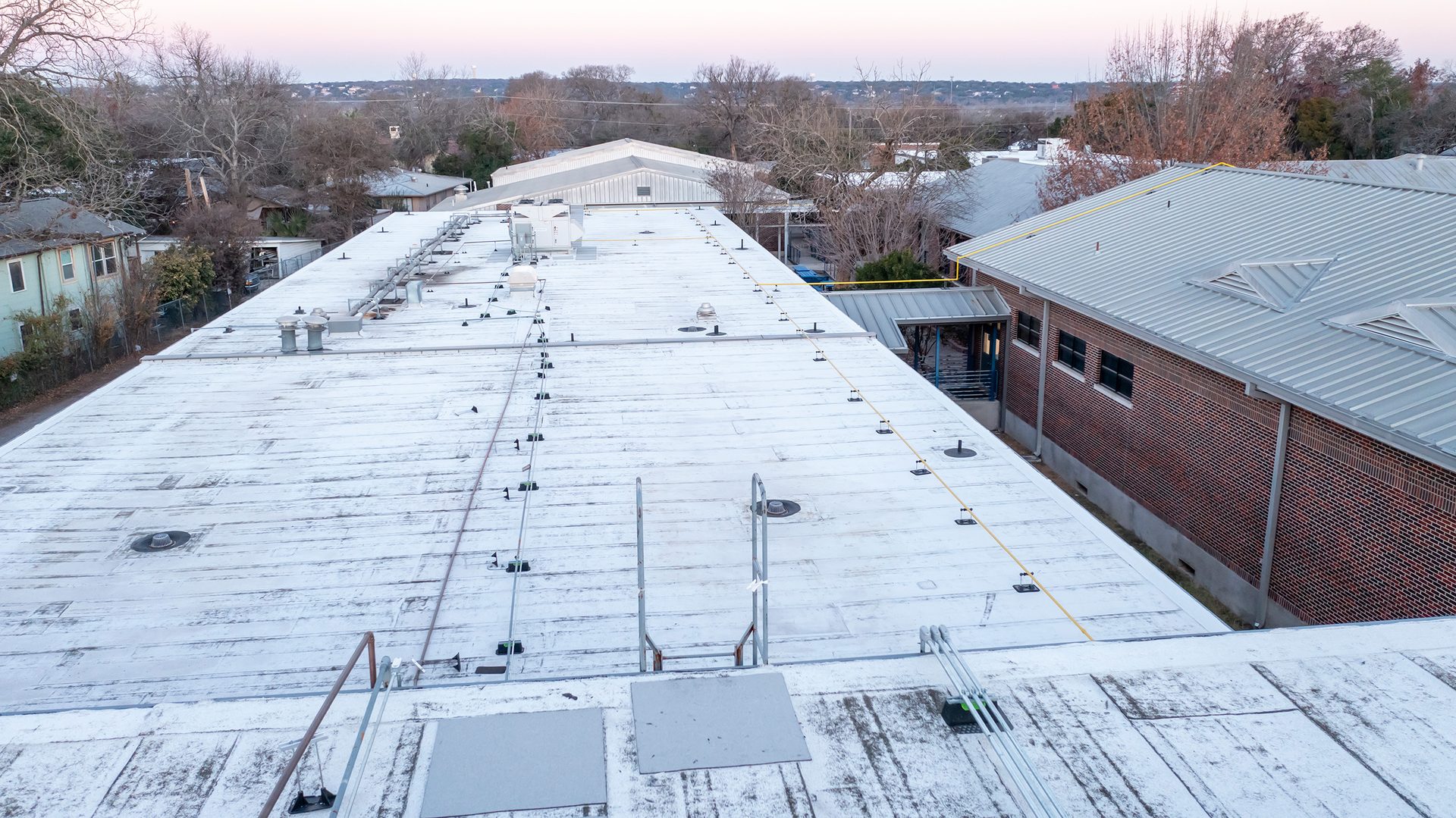 Aerial view of a white flat roof with equipment, adjacent to a brick building with a metal roof.