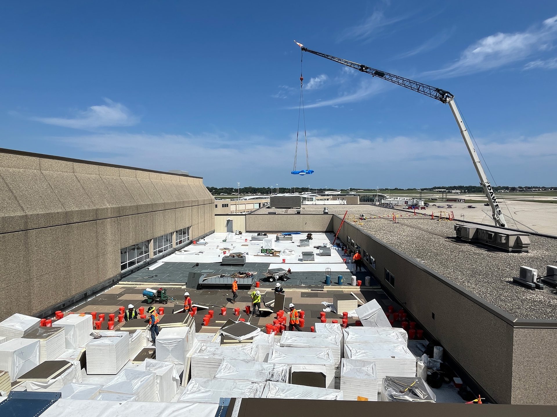 Crane lifts blue cargo onto a large roof where workers are preparing roofing materials at an airport.