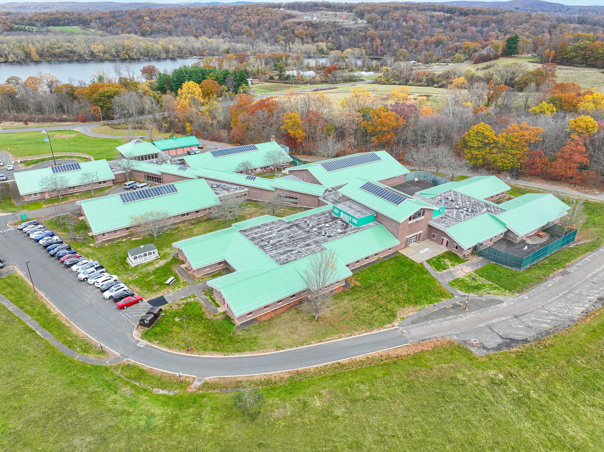 Drone view of a school with green roofs, solar panels, and parking lot, surrounded by autumn trees and a lake.