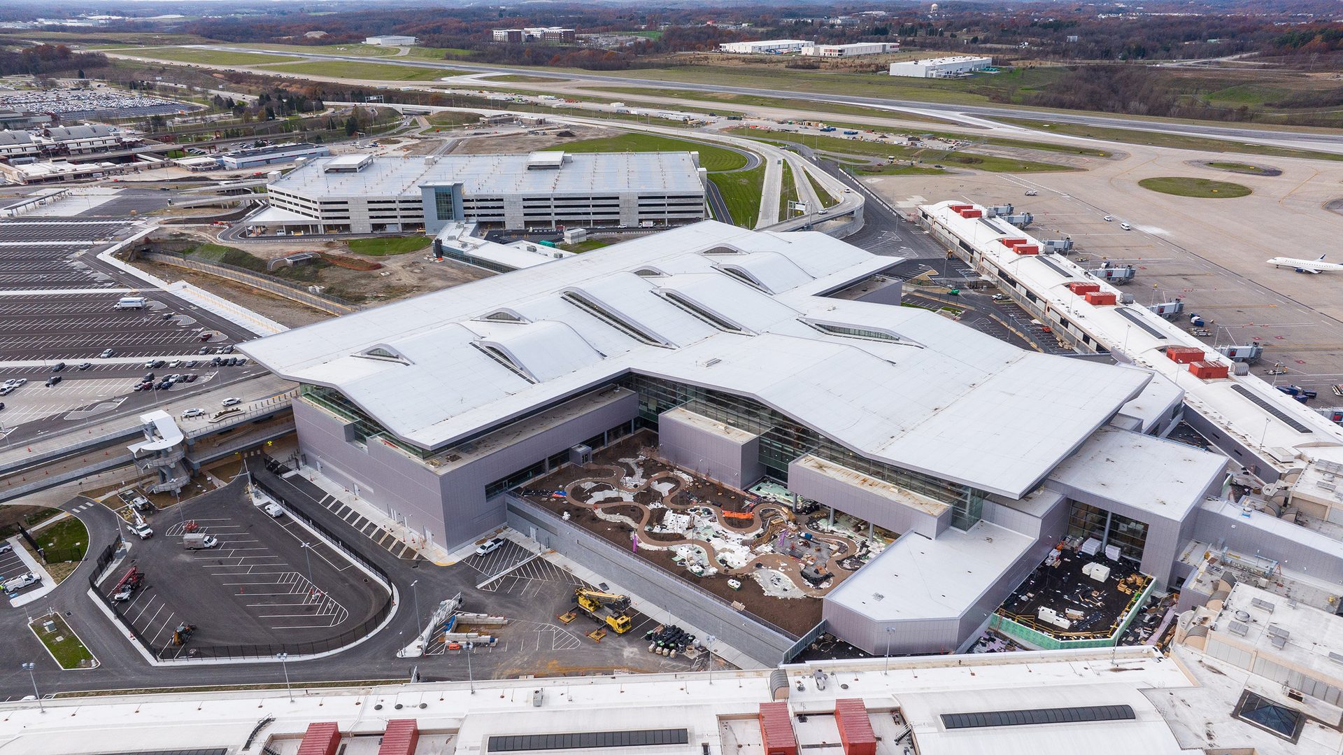 An aerial shot of a modern airport terminal building with a unique wavy roof, parking structures, and planes.