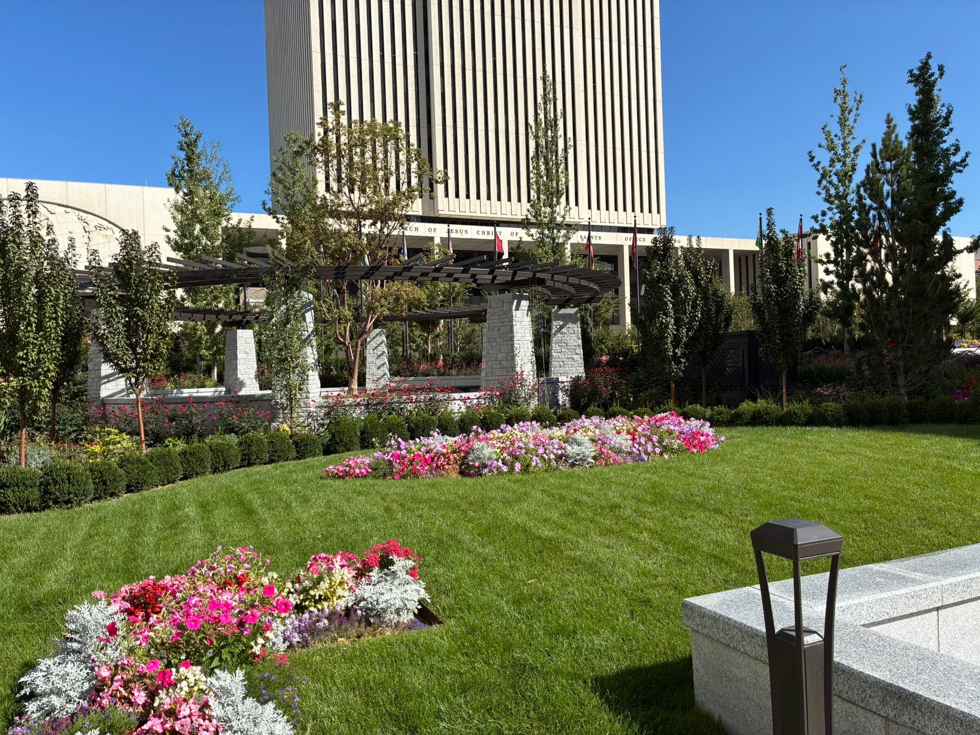 A vibrant garden with flower beds, a green lawn, and trees in front of a modern high-rise building on a sunny day.
