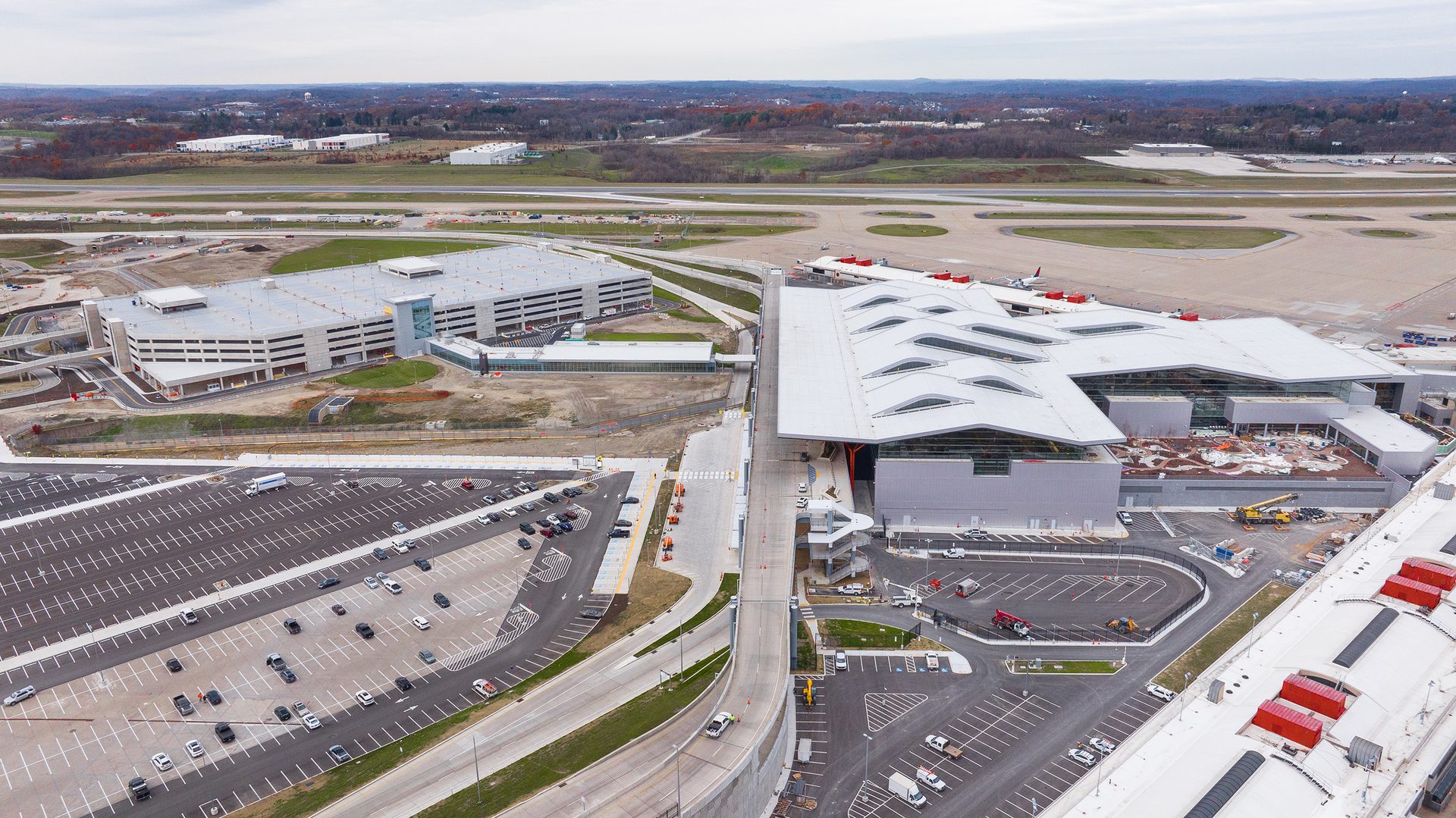 An aerial view showing a modern airport terminal, a large parking garage, and an open-air parking lot.