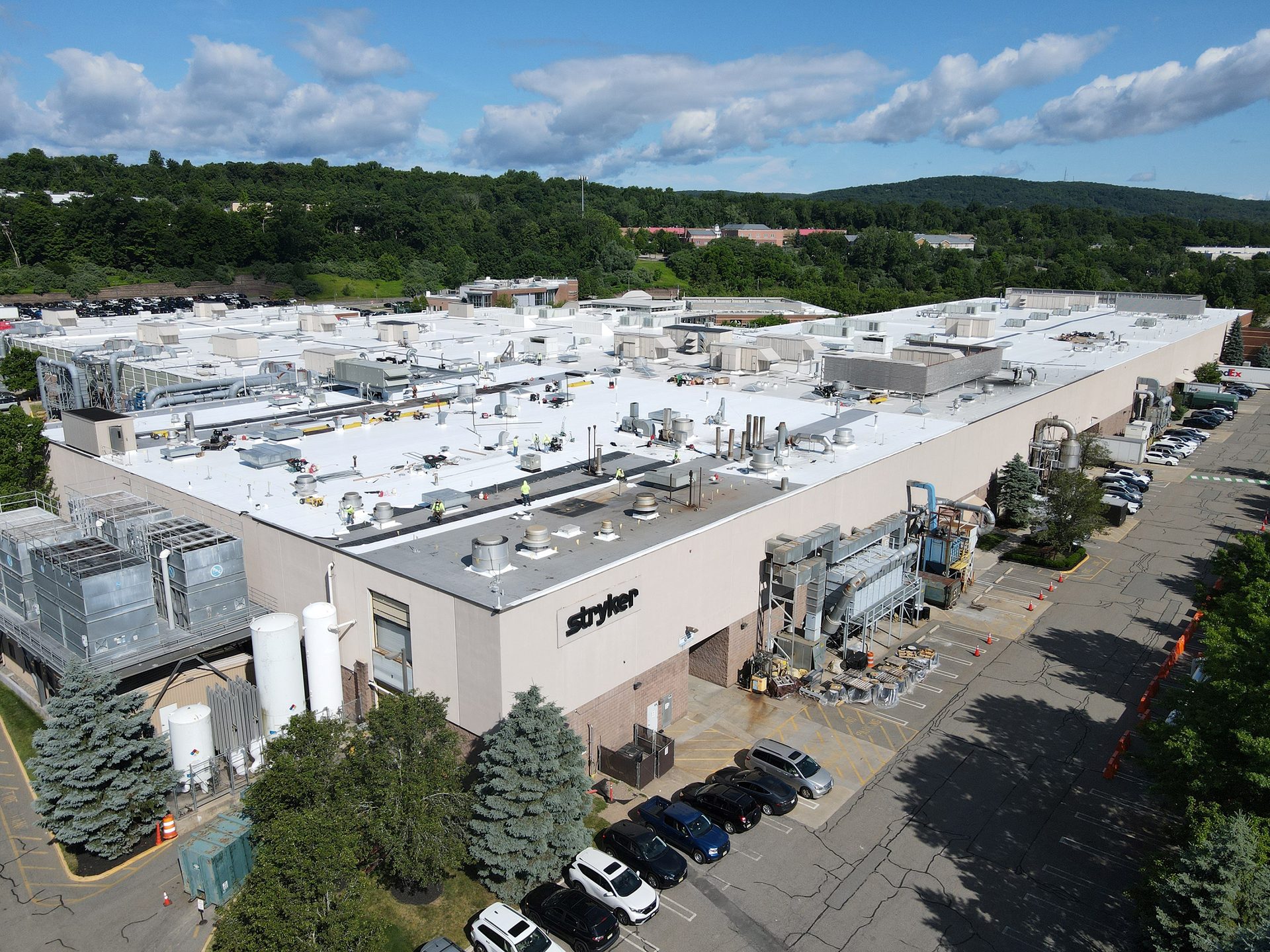 Aerial view of a large Stryker factory with white roof, industrial equipment, parking lots, and trees.