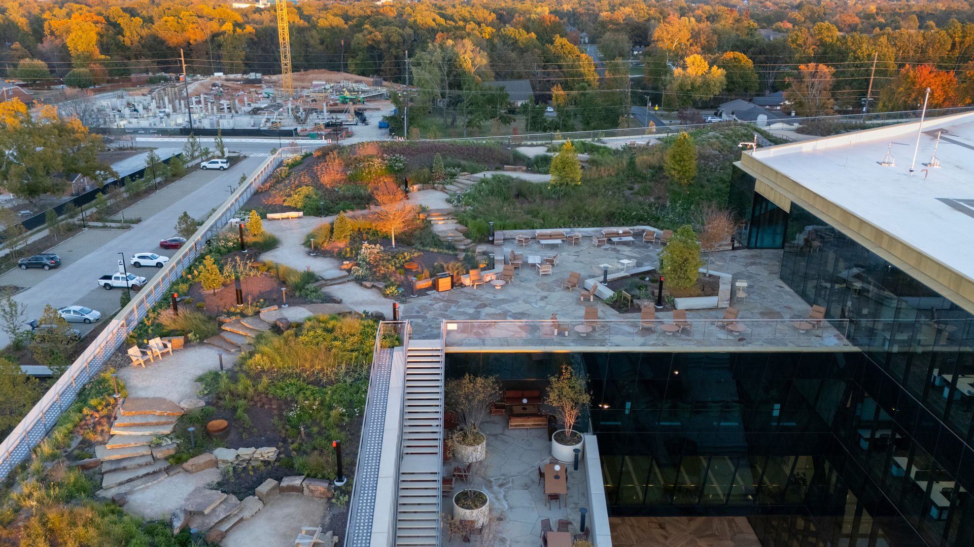 Aerial view of a modern building with a multi-level rooftop garden and patio seating, near a construction site.
