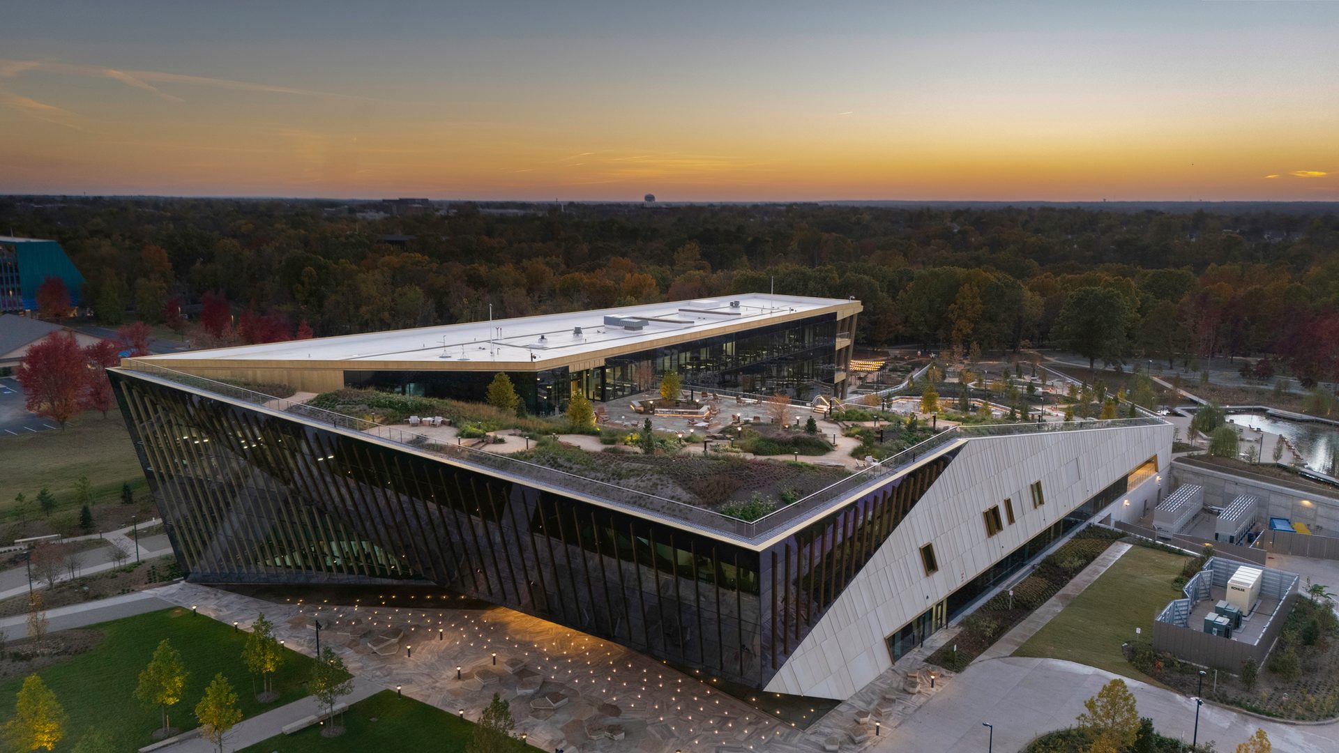Aerial view of a modern building with a large green roof and outdoor patio, surrounded by autumn trees at sunset.