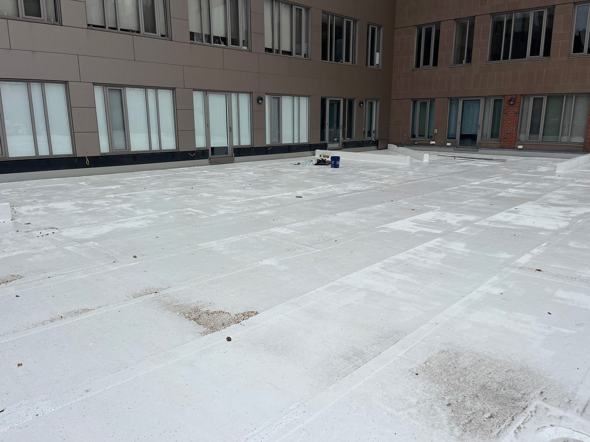 Wide view of a flat white rooftop patio with dirt patches, surrounded by a building with many windows.