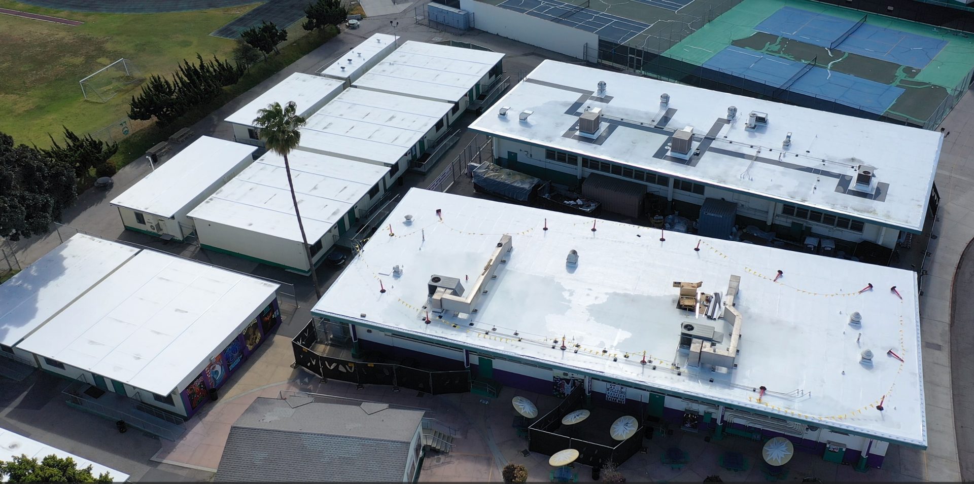 Aerial view of a school campus with several white-roofed modular buildings, a tall palm tree, and sports fields.