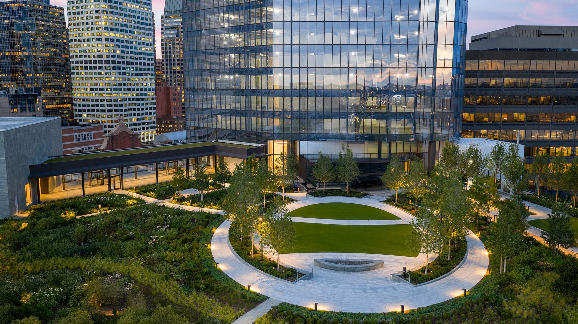 Aerial view of a modern city park with a circular lawn, trees, curved pathways, and skyscrapers at dusk.