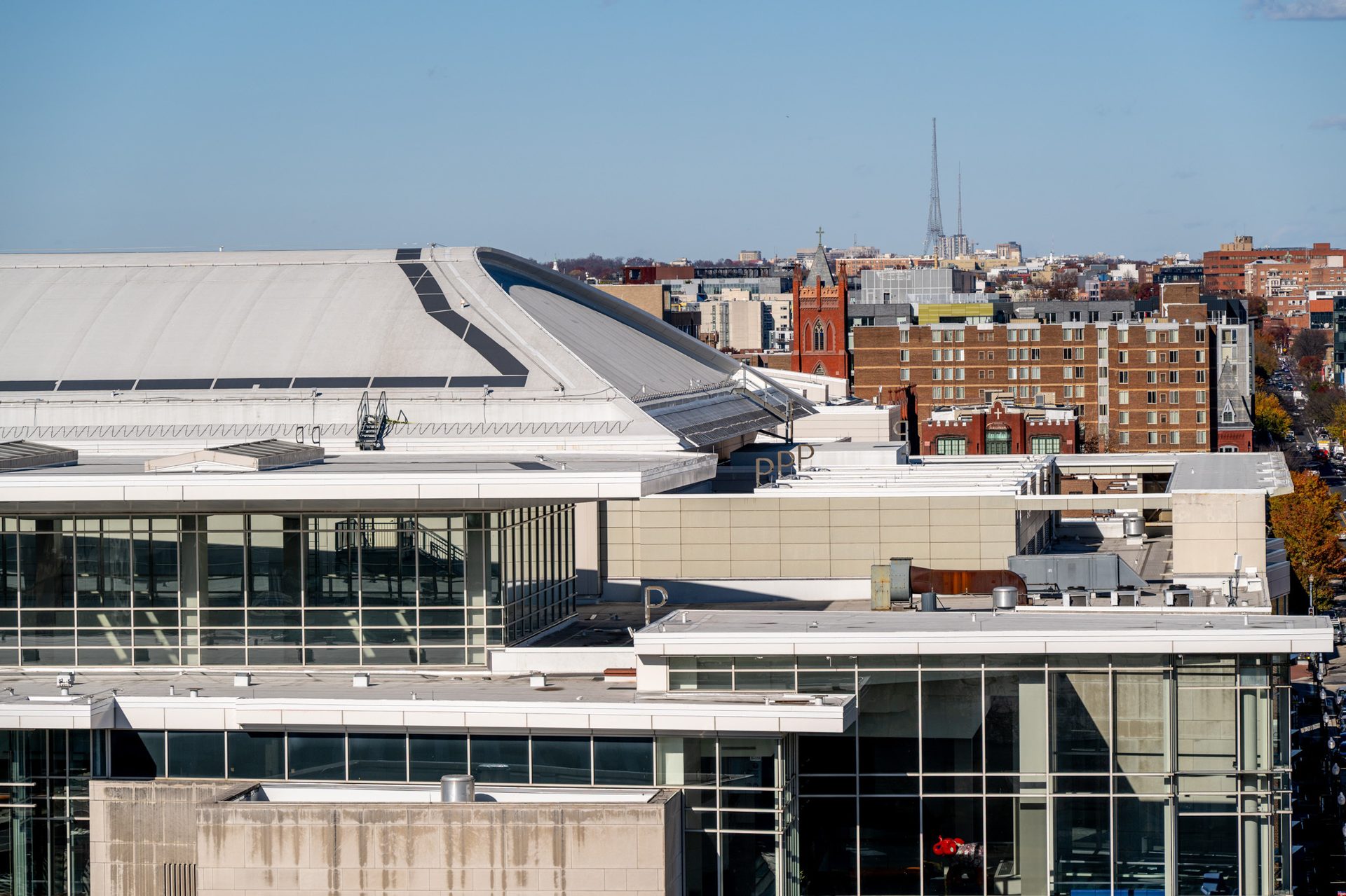 Aerial view of city buildings under a clear sky, featuring a large structure with a curved roof and distant towers.