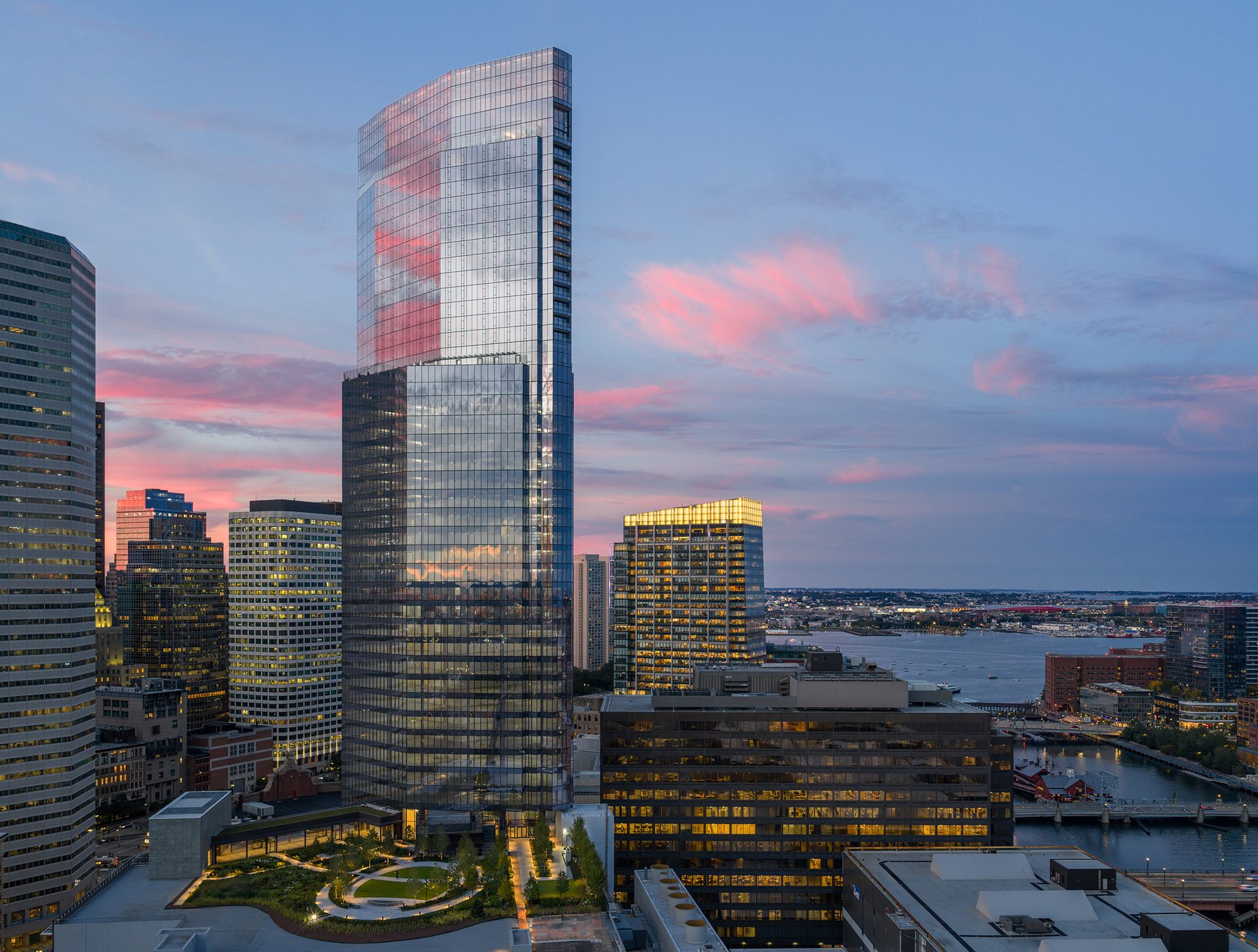 Aerial view of a modern glass skyscraper, rooftop garden, and city waterfront with a pink sunset sky.