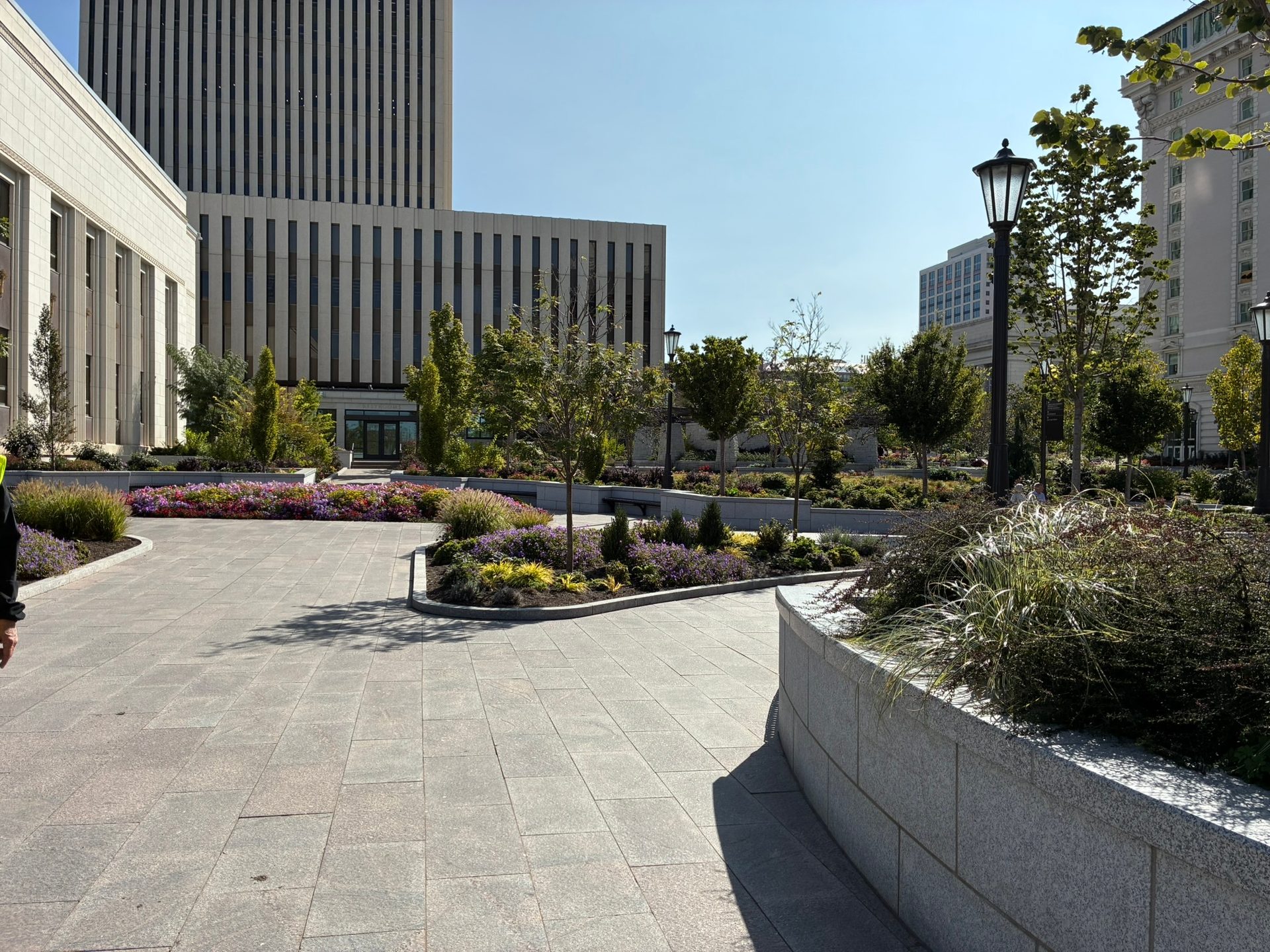 An urban plaza with modern buildings, paved walkways, and vibrant garden beds under a clear sky.