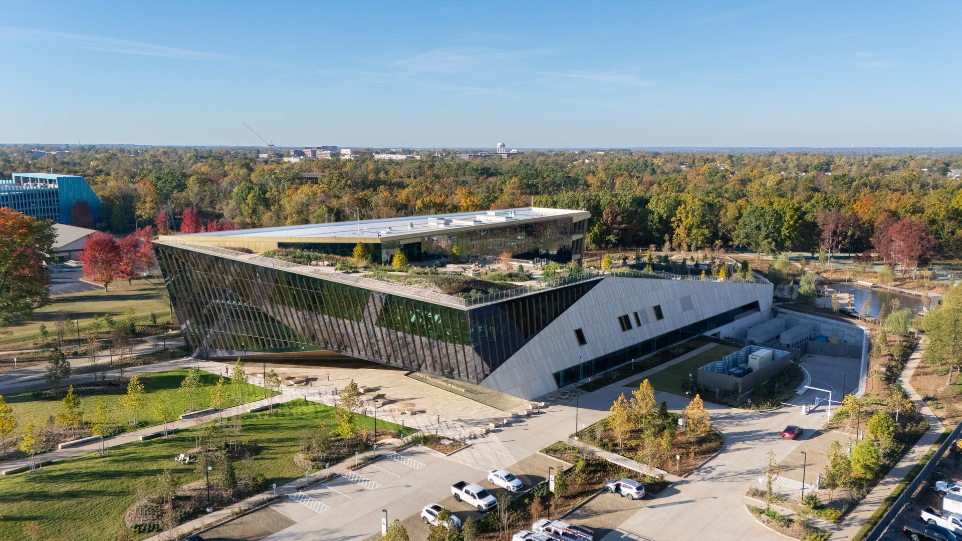 Aerial view of a modern angular building with a green roof, surrounded by autumn trees and a parking lot.