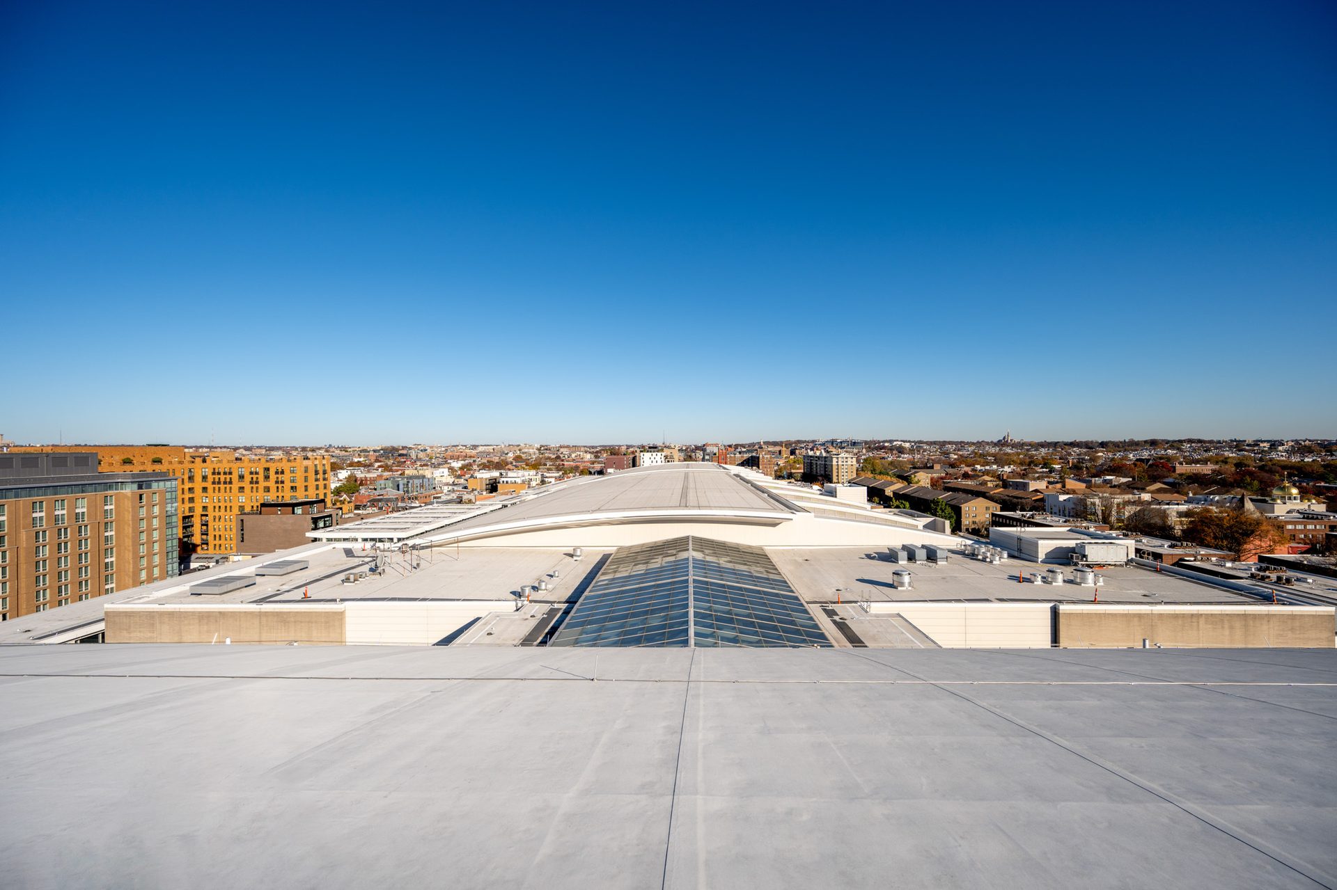 Aerial view of a large white roof with a glass skylight, backed by a sprawling city skyline under a clear blue sky.