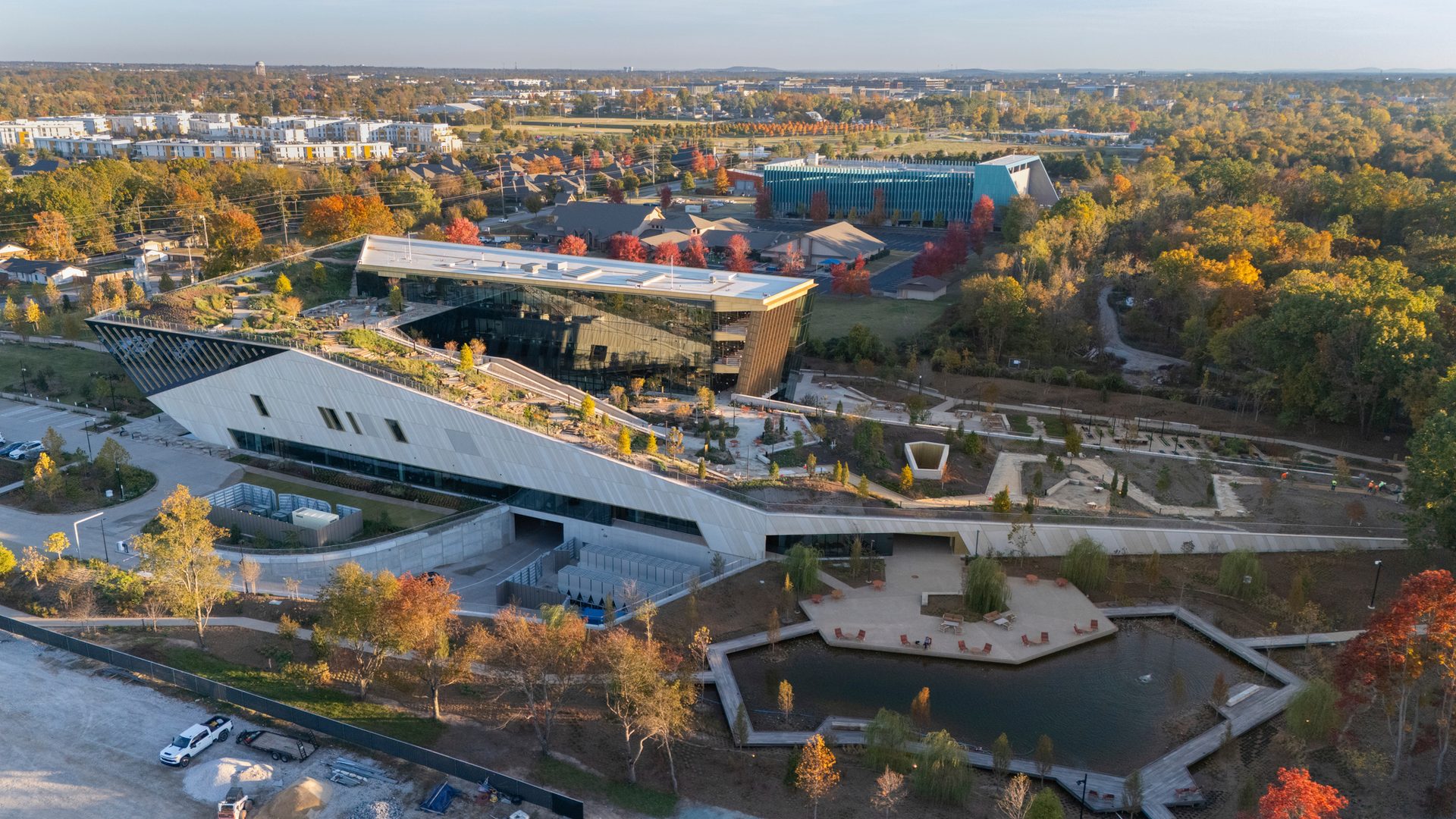 Aerial view of a modern building with a green roof, glass facade, pond, and autumn foliage.