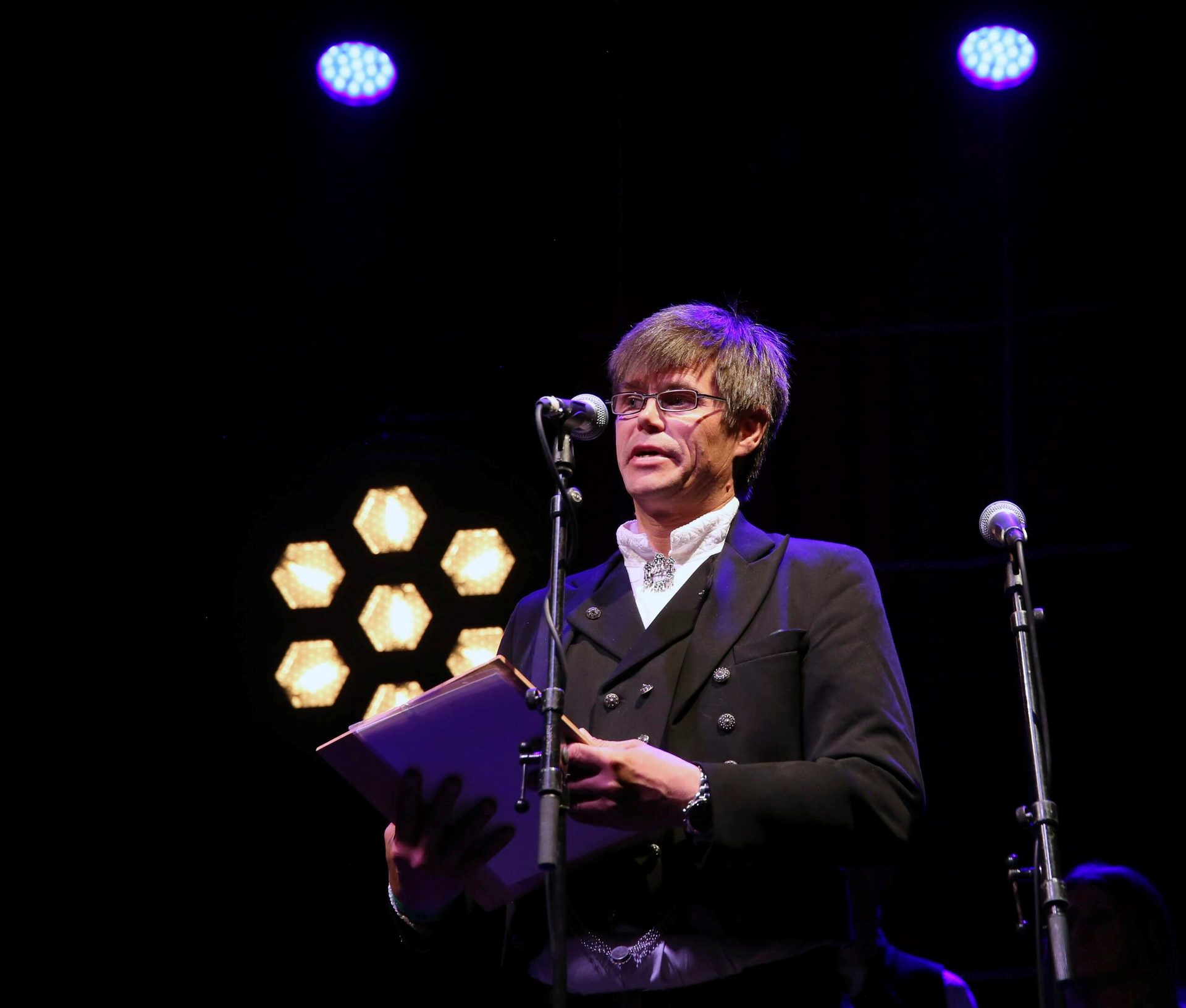 Man in glasses speaks into mic, holding a book, on a dark stage with glowing lights.