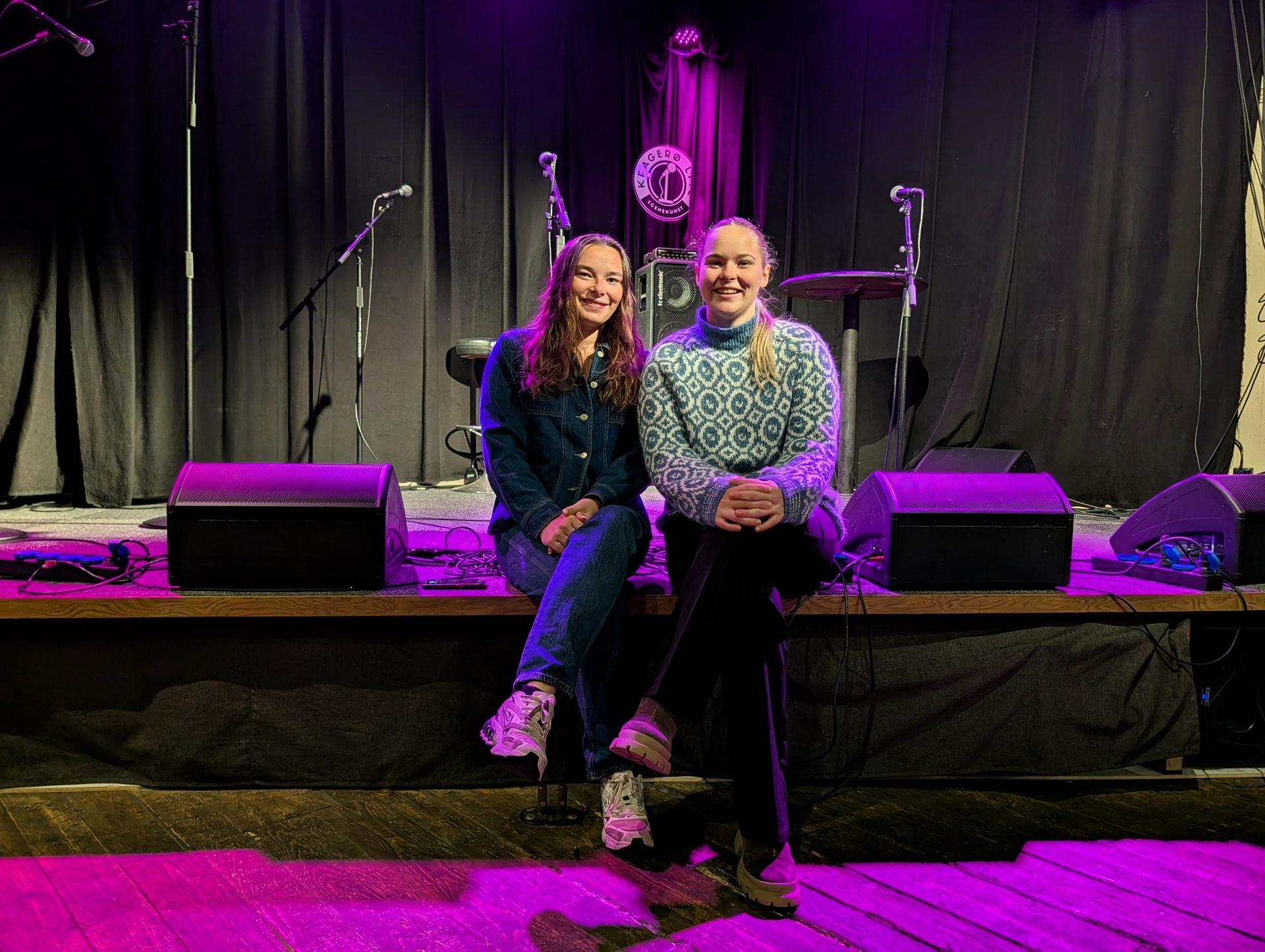 Two smiling women sit on a stage with microphones and purple lighting.