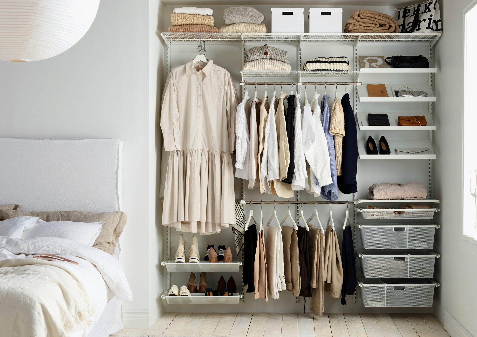 An open white closet system filled with neutral-toned clothes, shoes, and accessories, next to a bed.