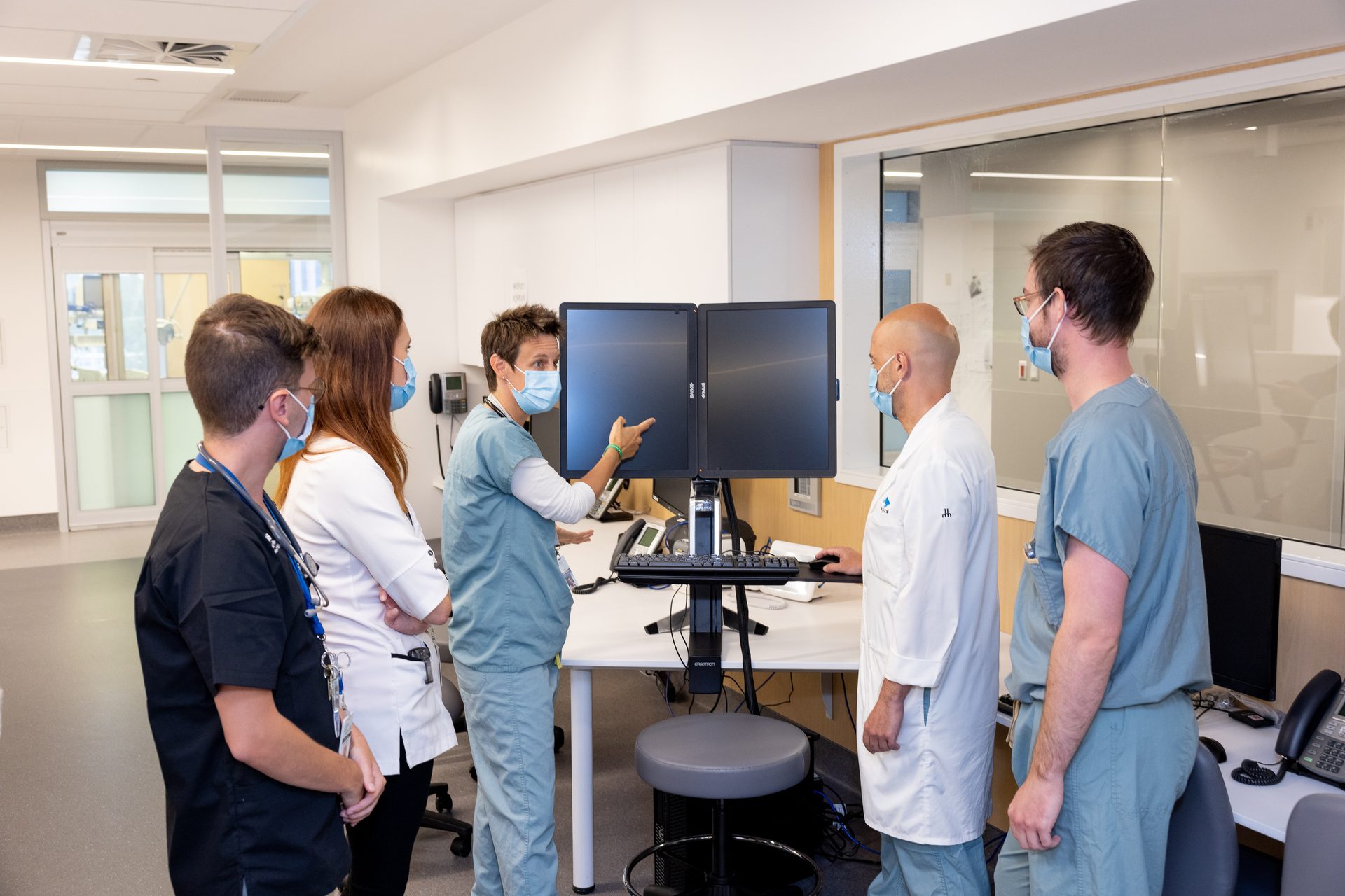 Medical staff in masks reviewing data on dual monitors in a clinic, with one person pointing.