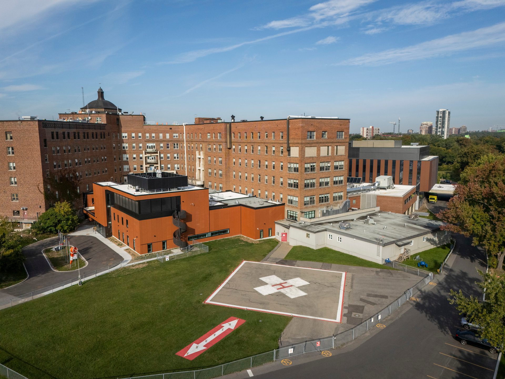 An aerial view shows a large brick hospital complex with a modern annex, green lawn, and helipad.