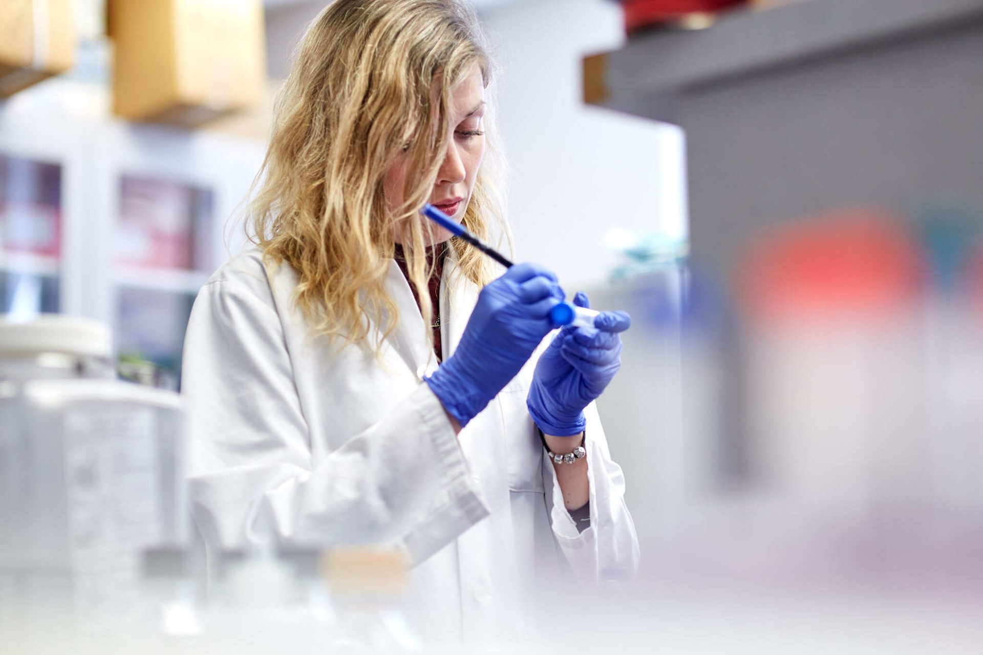 A female scientist in a lab coat and blue gloves carefully pipetting into a small vial in a lab.