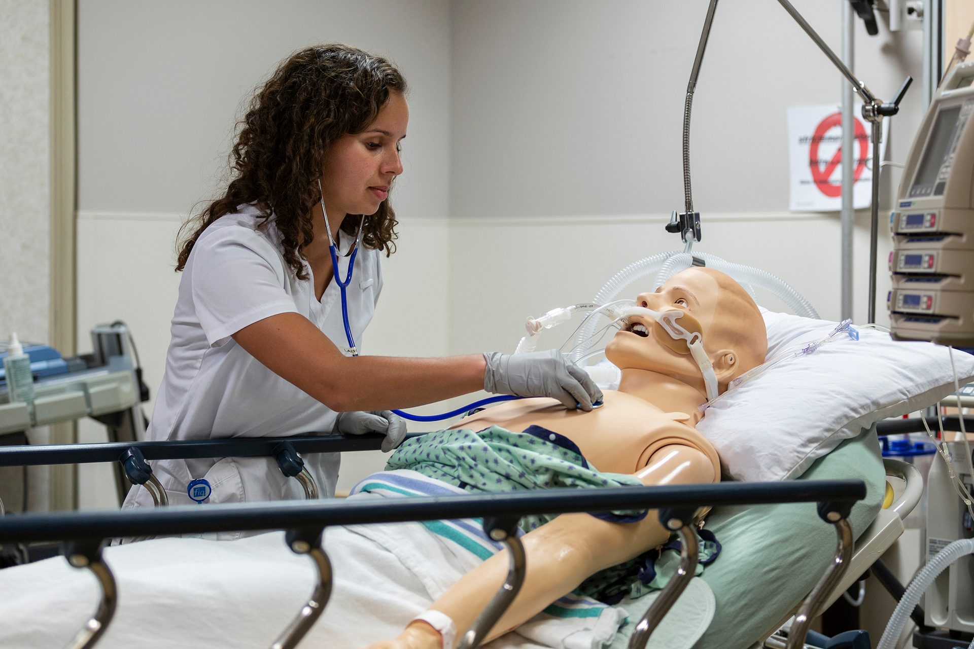 A nurse uses a stethoscope on a medical training dummy lying on a bed with ventilator tubes.