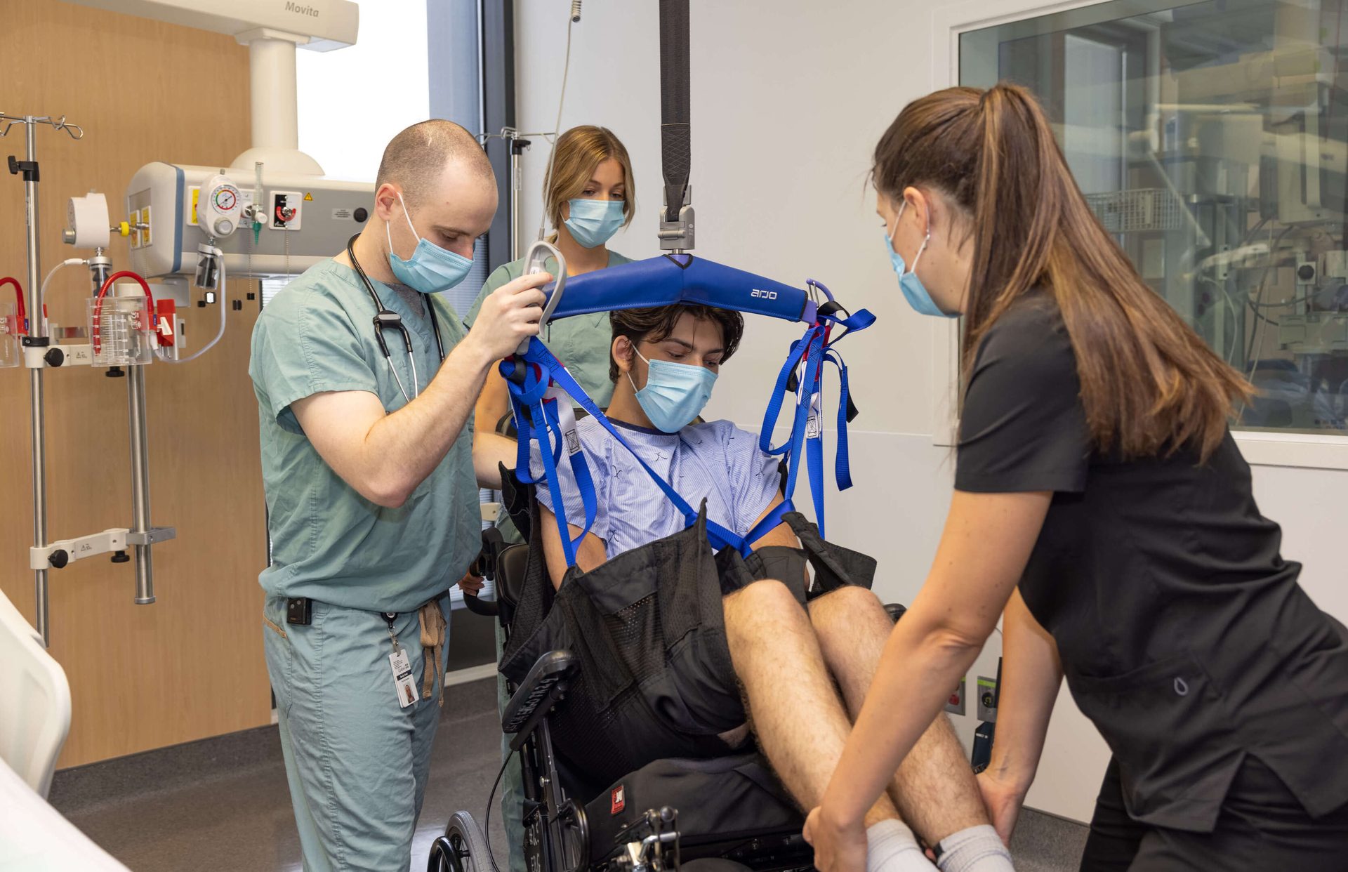 Three healthcare workers assist a masked patient being transferred from a wheelchair using a lift harness.