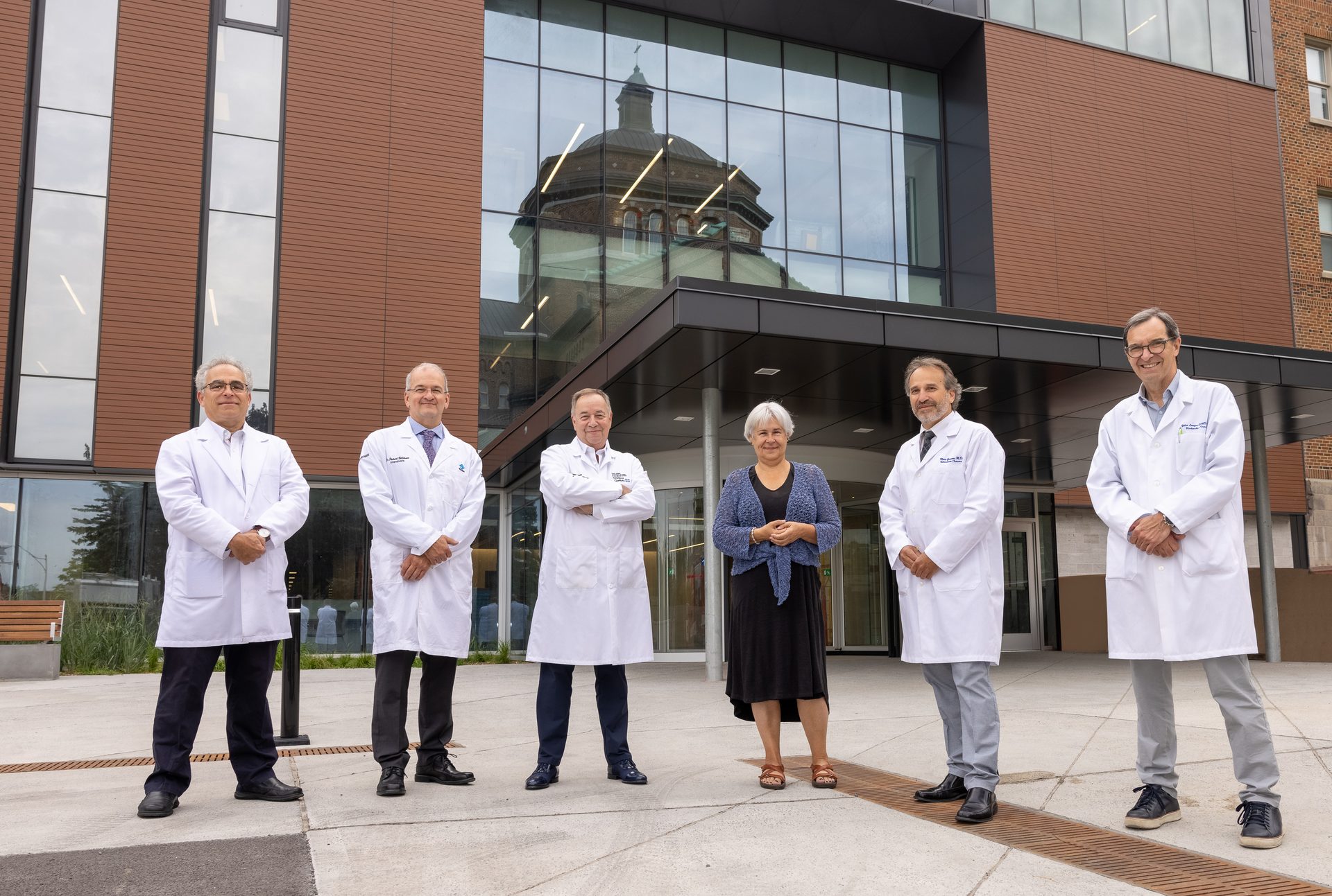 Six medical professionals, five men and one woman, stand in front of a modern building with glass reflections.