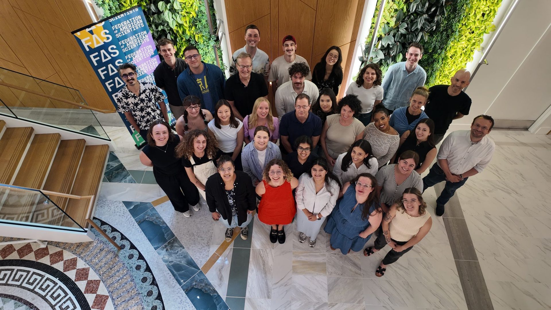 A large, diverse group of smiling people looking up, posed on tiered steps with a "FAS" banner.