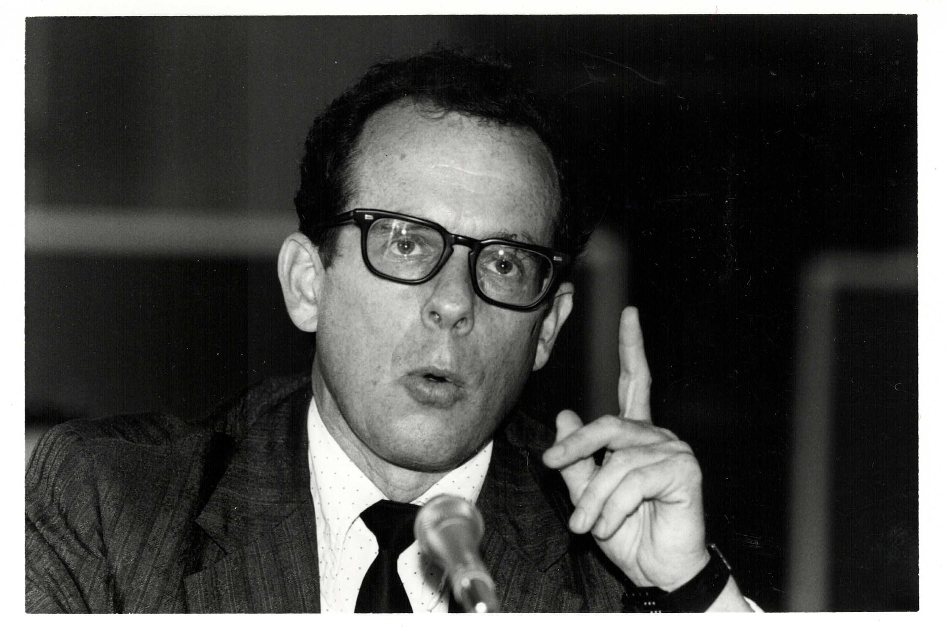 Black and white photo of a man in glasses speaking, gesturing with a raised finger.