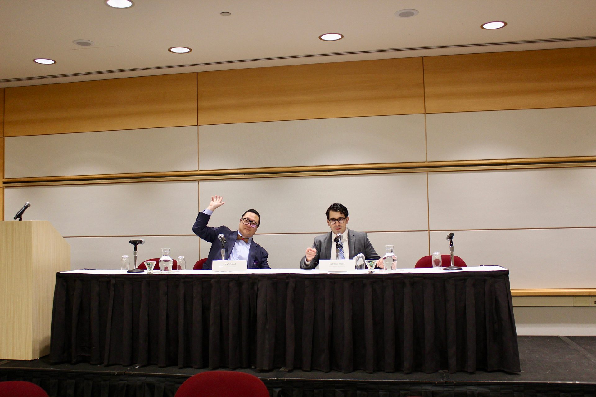 Two men sit at a conference panel table; one waves, the other gestures.