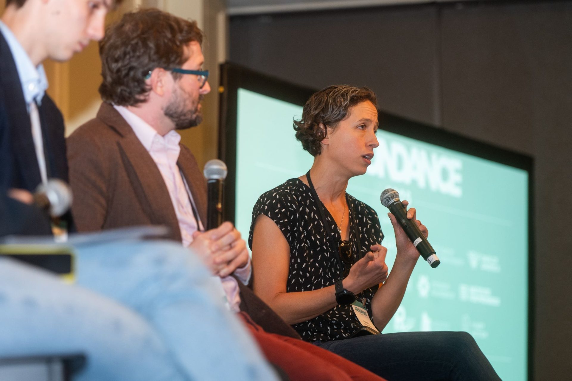 Woman speaking into a microphone at a panel with others and a screen.