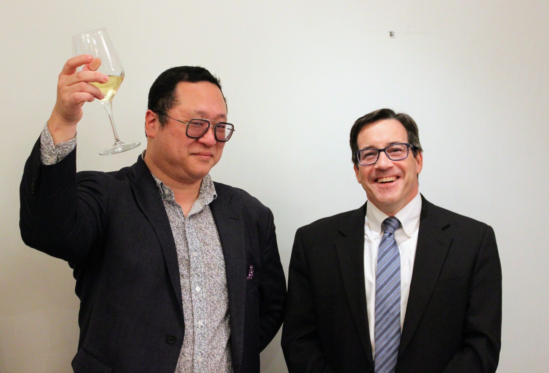 Two smiling men, one raising a glass of white wine.