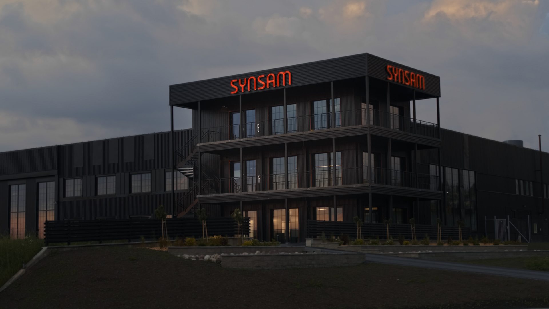 A modern, dark building with a glowing red "SYNSAM" sign under a cloudy sky.