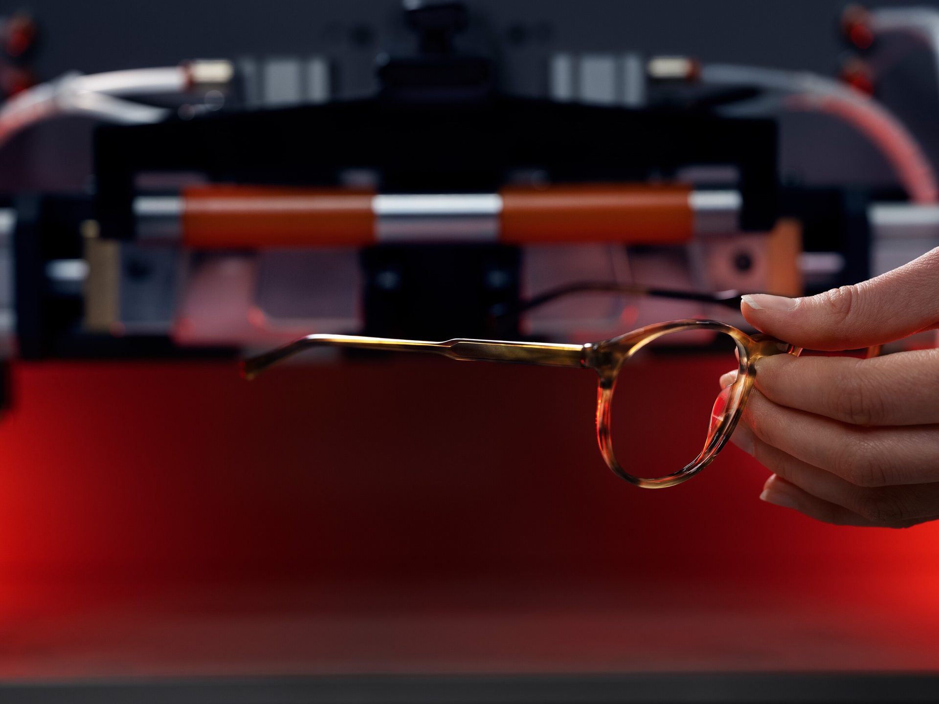 Hand holds tortoiseshell eyeglasses against a red-lit, blurry industrial background.