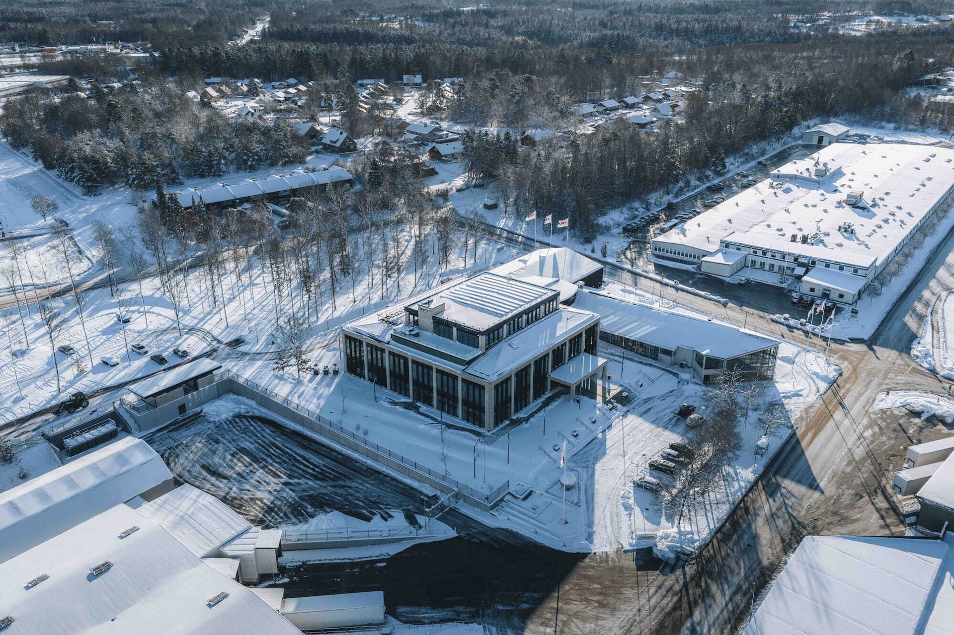 Aerial view of snow-covered modern buildings and a large factory, surrounded by winter trees and roads.