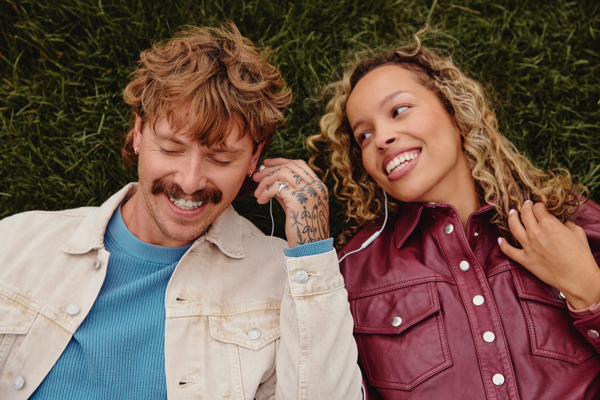 A man and woman lie on grass, smiling and sharing earbuds.