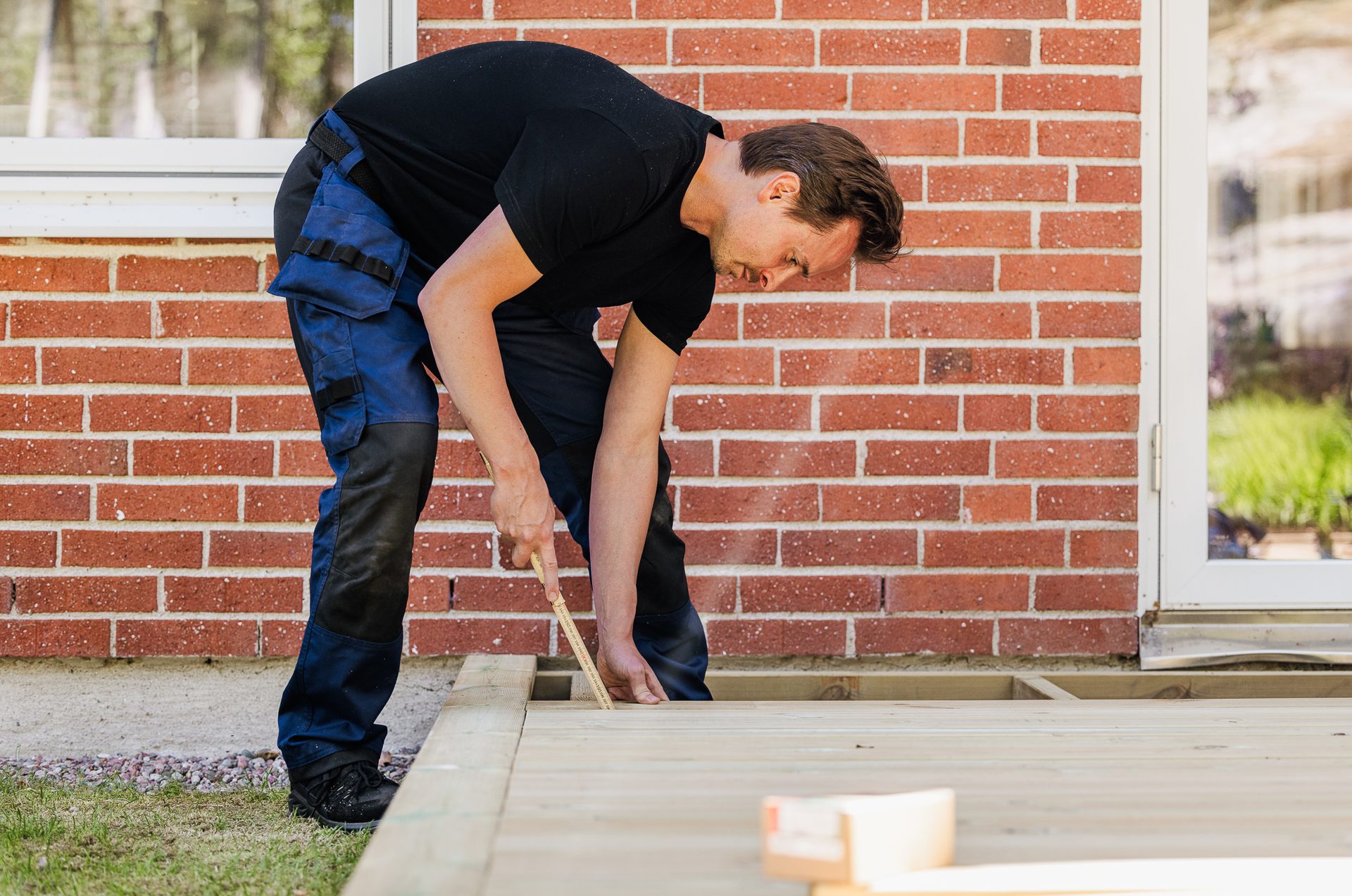 Man in work pants and black shirt measuring and working on a wooden deck next to a brick wall.