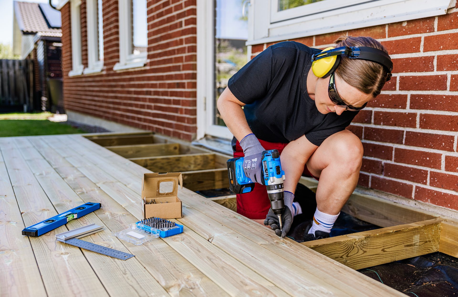 Person in safety gear drilling a screw into a wooden deck board during construction.