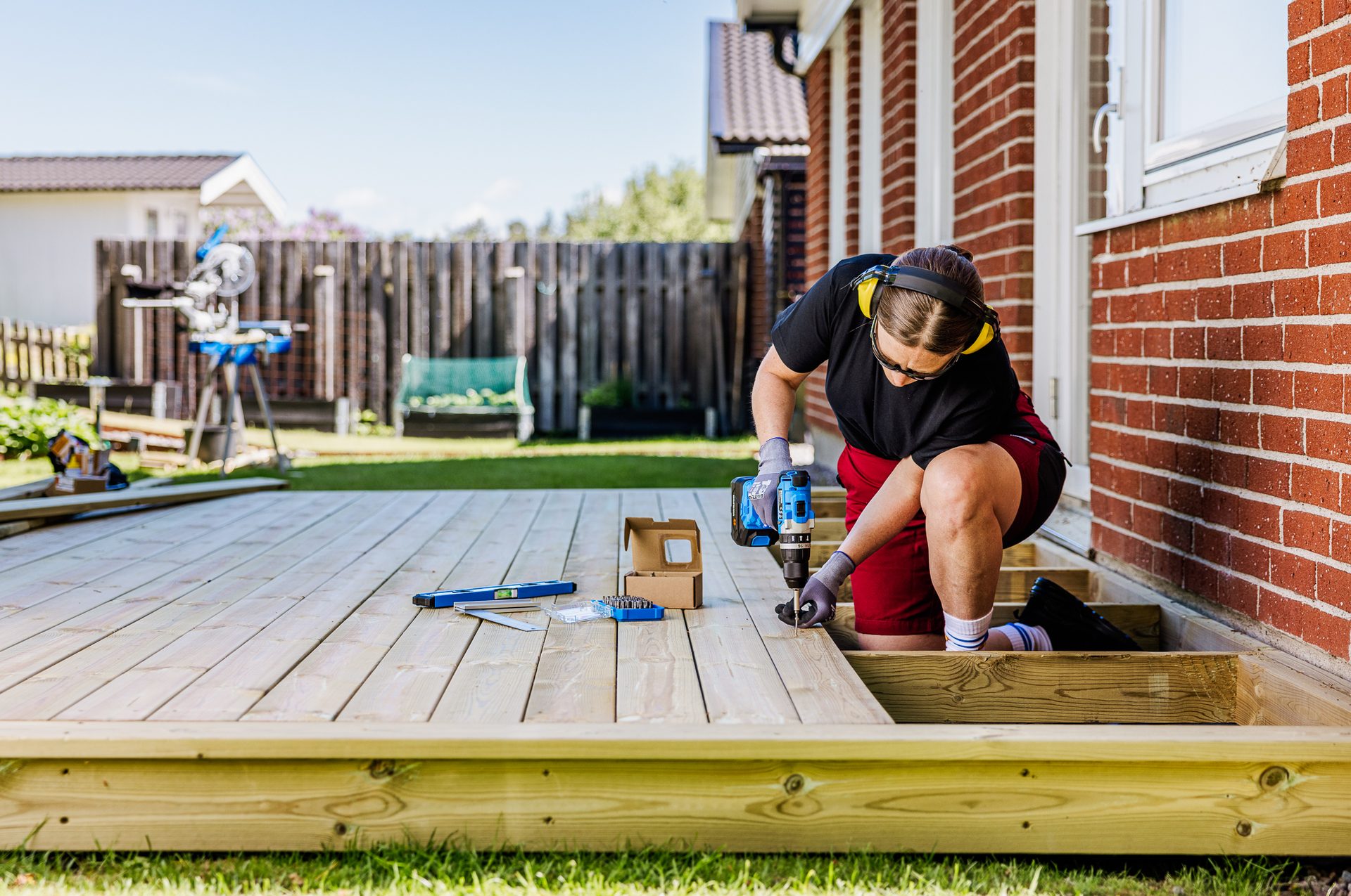 Woman installing deck boards with a power drill in a sunny backyard. She wears safety goggles and ear protection.