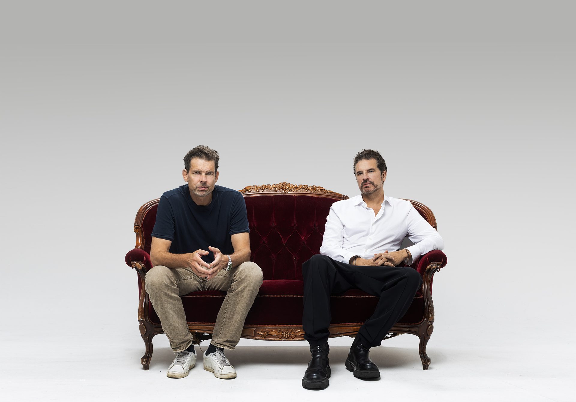 Two men sit on a red velvet couch, looking at the camera.