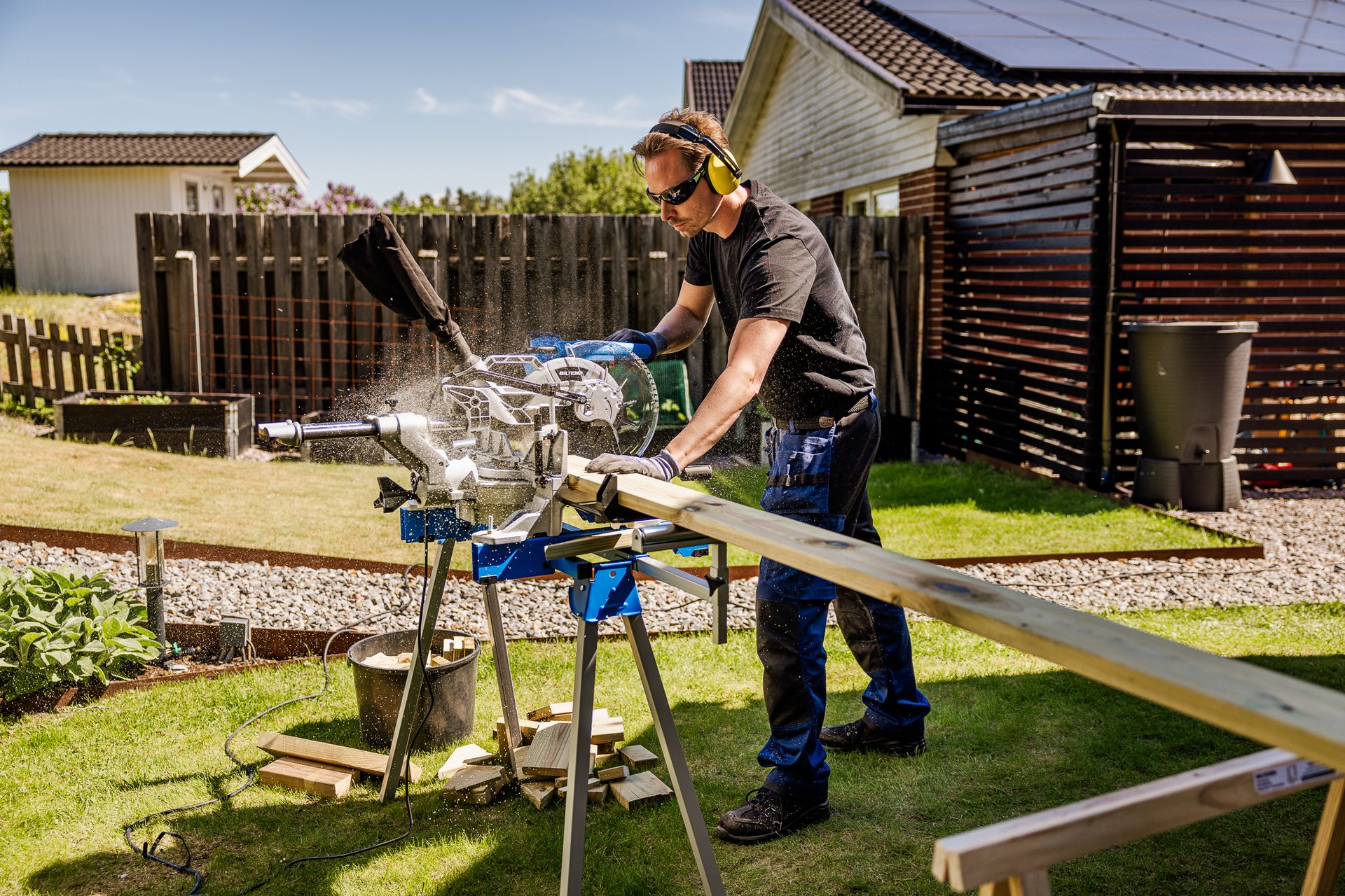 Man wearing safety glasses and ear protection cuts wood with a miter saw outdoors, creating sawdust.