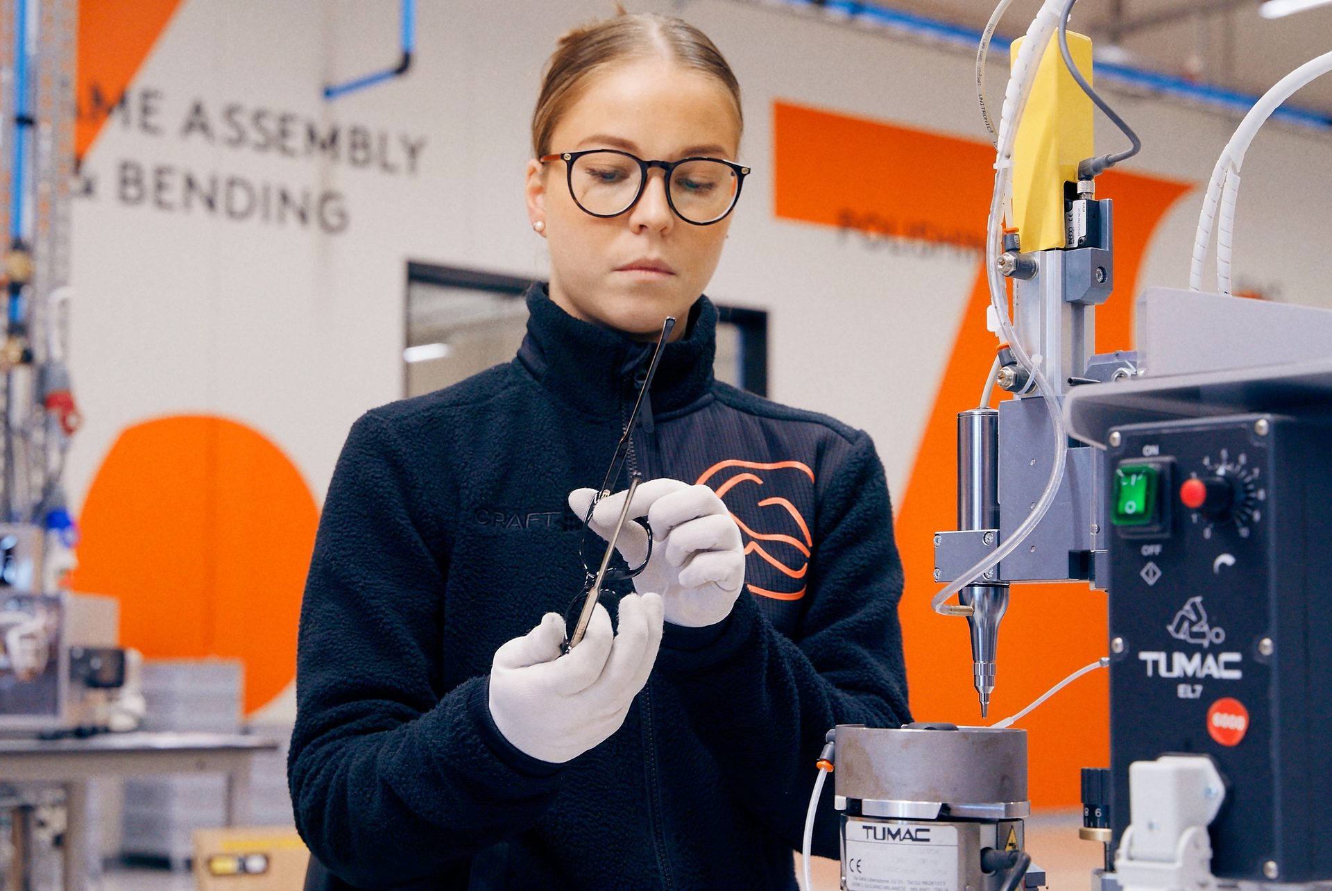Woman in glasses and gloves assembling eyewear at a TUMAC machine.