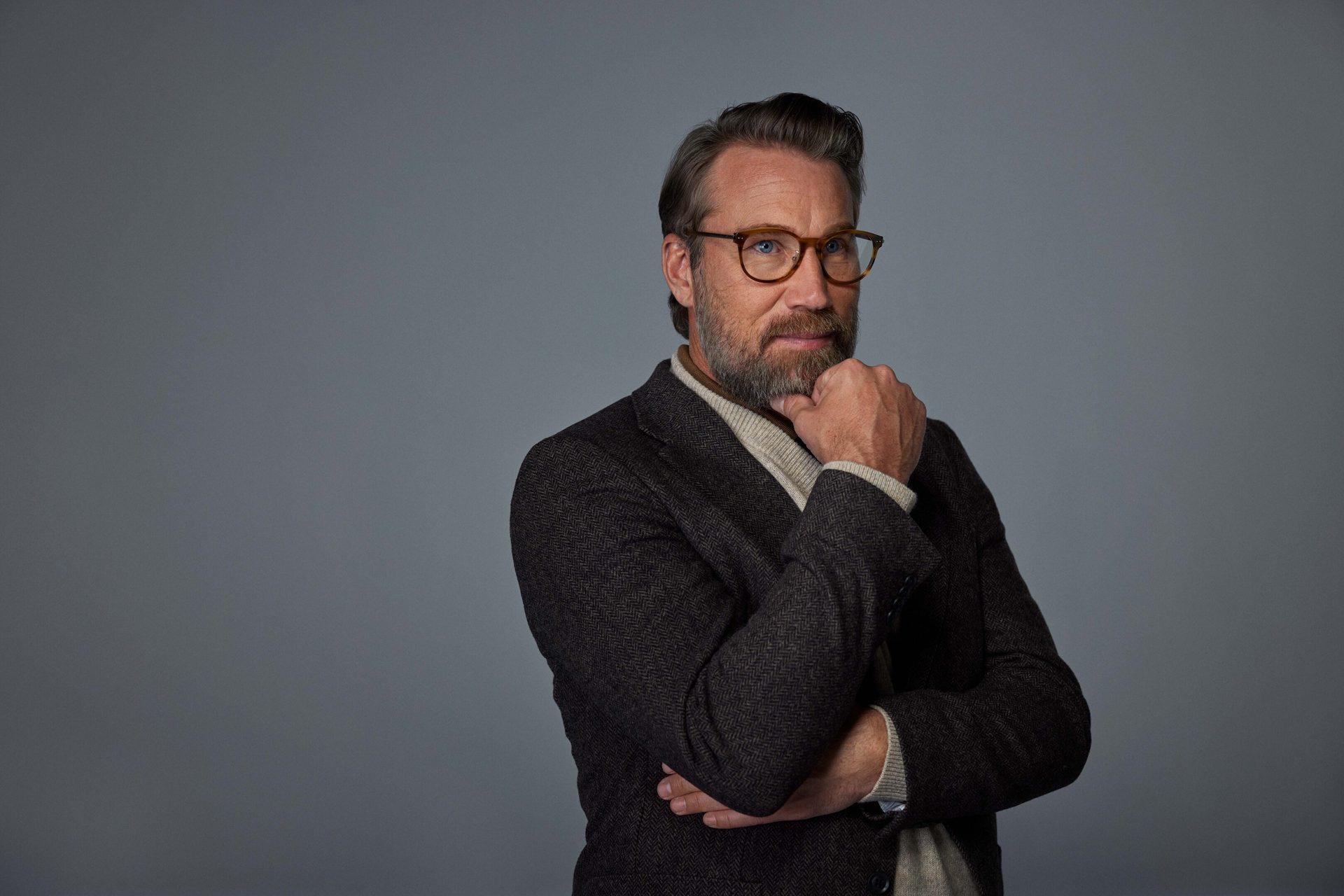 Pensive man with beard and glasses in tweed jacket, hand on chin, against gray backdrop.