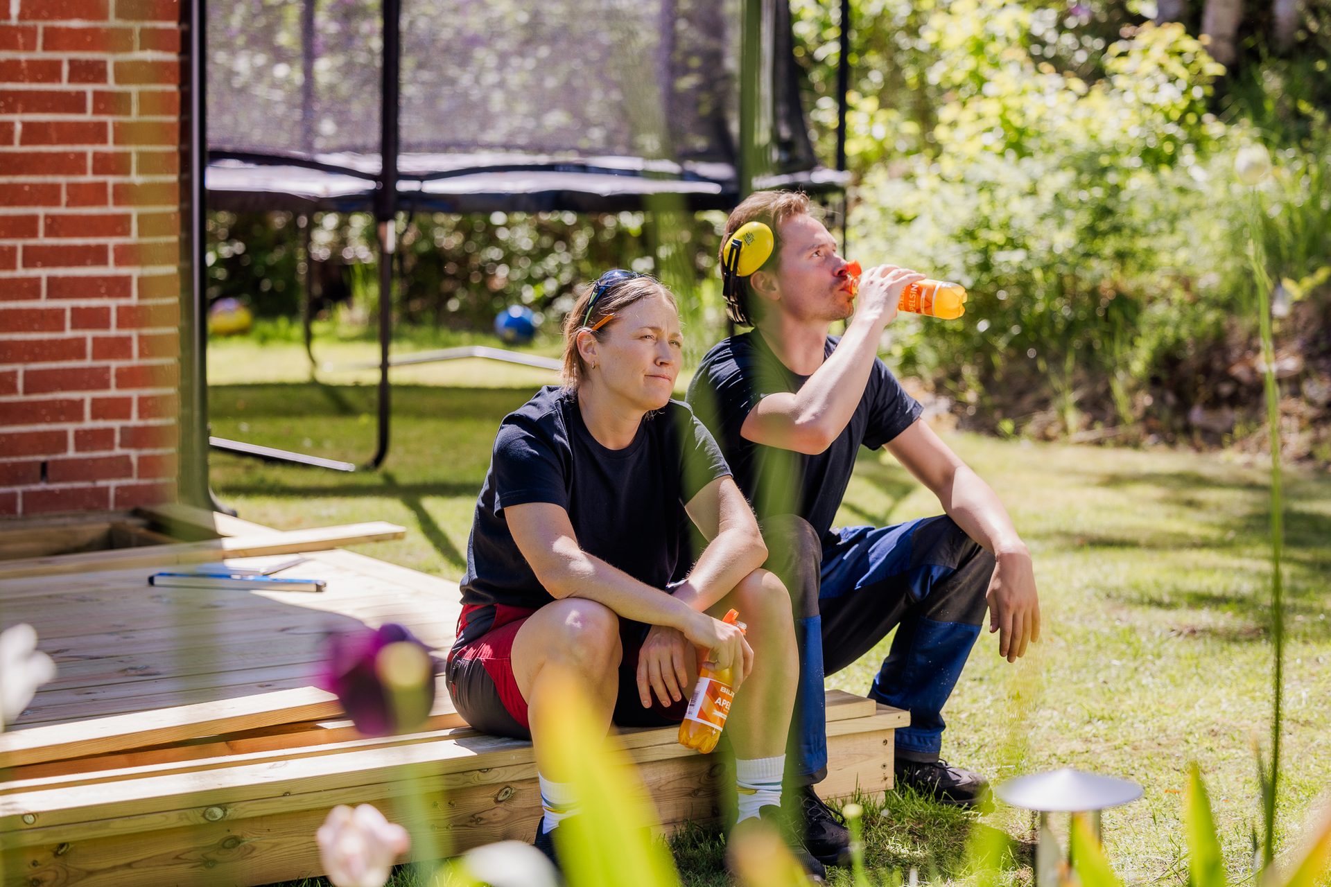 Two people, a man and a woman, taking a break on a wooden deck, drinking from orange bottles on a sunny day.