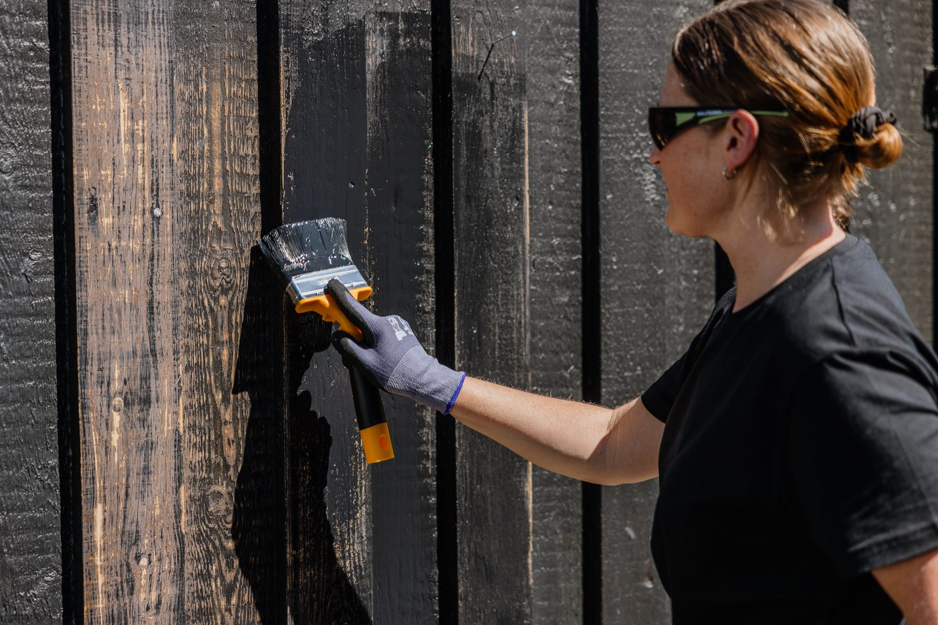 A person in sunglasses and gloves paints a wooden fence black with a brush.